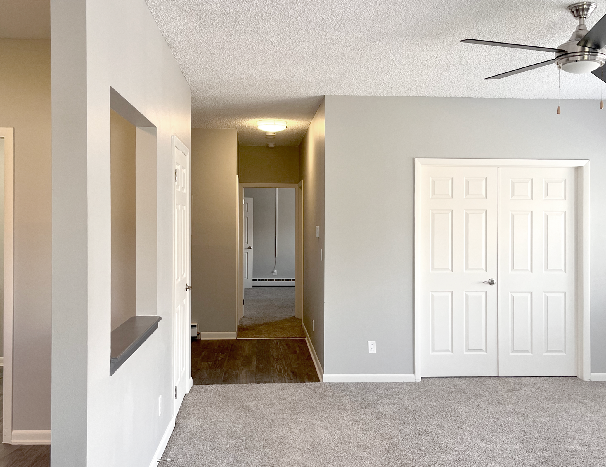 Interior view of a light gray living space featuring a doorway leading to a hallway, a ceiling fan, and two white doors. The walls are painted gray and the floors are carpeted. There is an open arch leading to a small nook, and another door on the right side, suggesting a simple and modern layout.