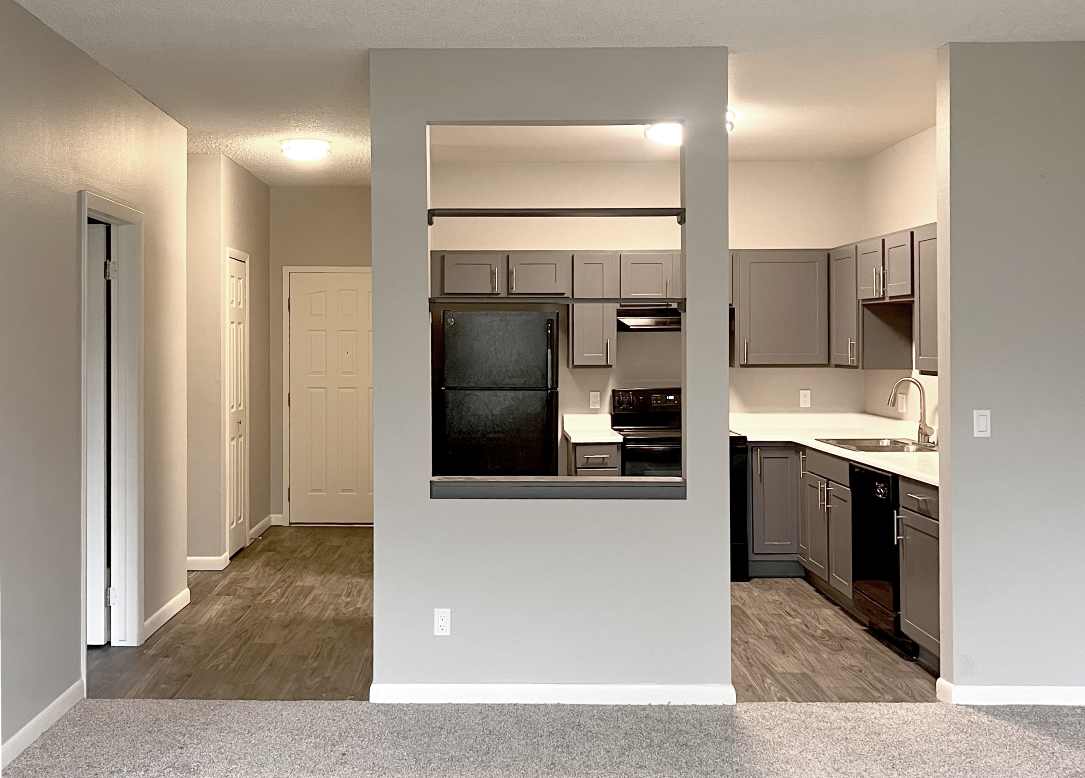 An open-concept living area featuring a modern kitchen with gray cabinets, black appliances, and a white countertop. The space is well-lit with natural light. The adjacent area includes a door leading to a hallway with additional doors, likely for rooms or storage, all with neutral-toned walls and flooring.