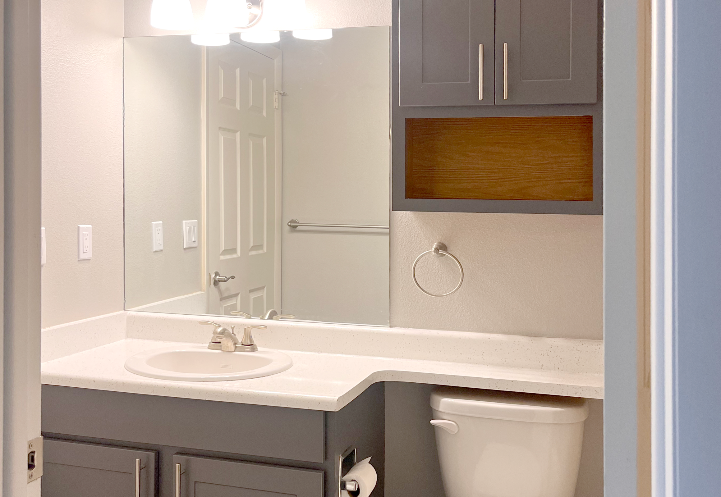 A modern bathroom featuring a gray vanity with a white countertop and sink. There is a large mirror above the sink, a light fixture with three bulbs, and a towel ring mounted on the wall. The bathroom includes a toilet and cabinetry with a wood accent shelf. The walls are painted in a light color, enhancing the space's brightness.