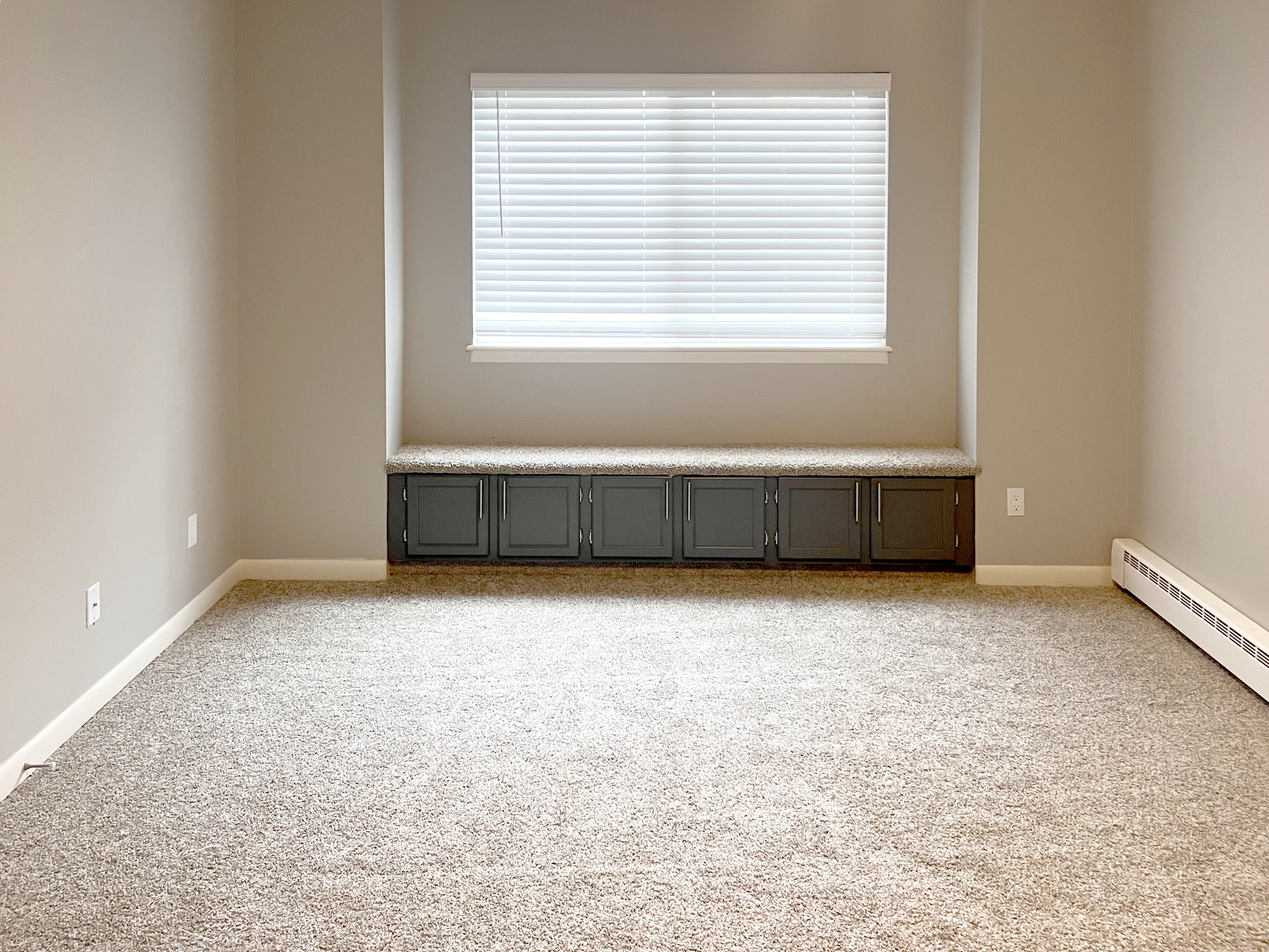 A spacious, empty room featuring light gray walls and a large window with white blinds. There is a built-in bench with storage underneath along one wall, and the floor is covered in soft, beige carpeting. The overall ambiance is clean and minimalistic, creating a bright and inviting space.