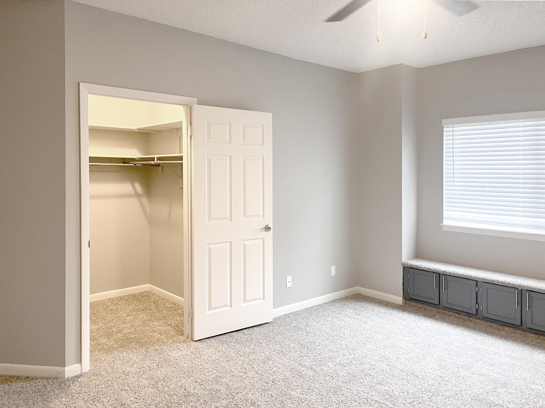 A cozy, well-lit bedroom featuring light gray walls and carpet. To the left, an open closet with shelves is visible through a white door, while a window on the right allows natural light to enter. A built-in seating area below the window adds charm to the space, creating a comfortable atmosphere.