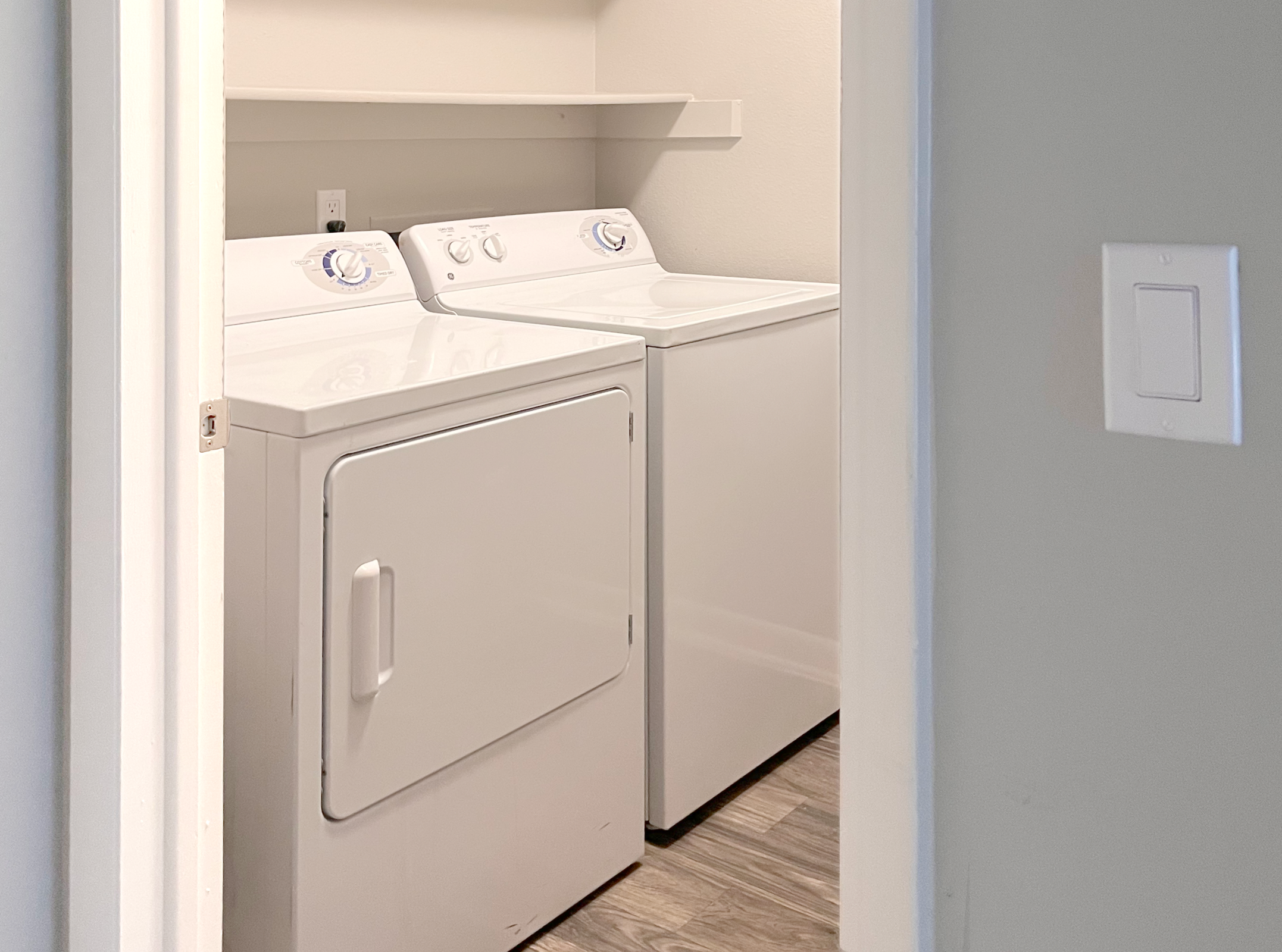 View of a small laundry room with a stacked washer and dryer unit. The appliances are white and located against a light-colored wall. A shelf is mounted above the washer and dryer, and the room has a clean, minimalistic appearance with wooden flooring. A door frame is visible on the left side of the image.