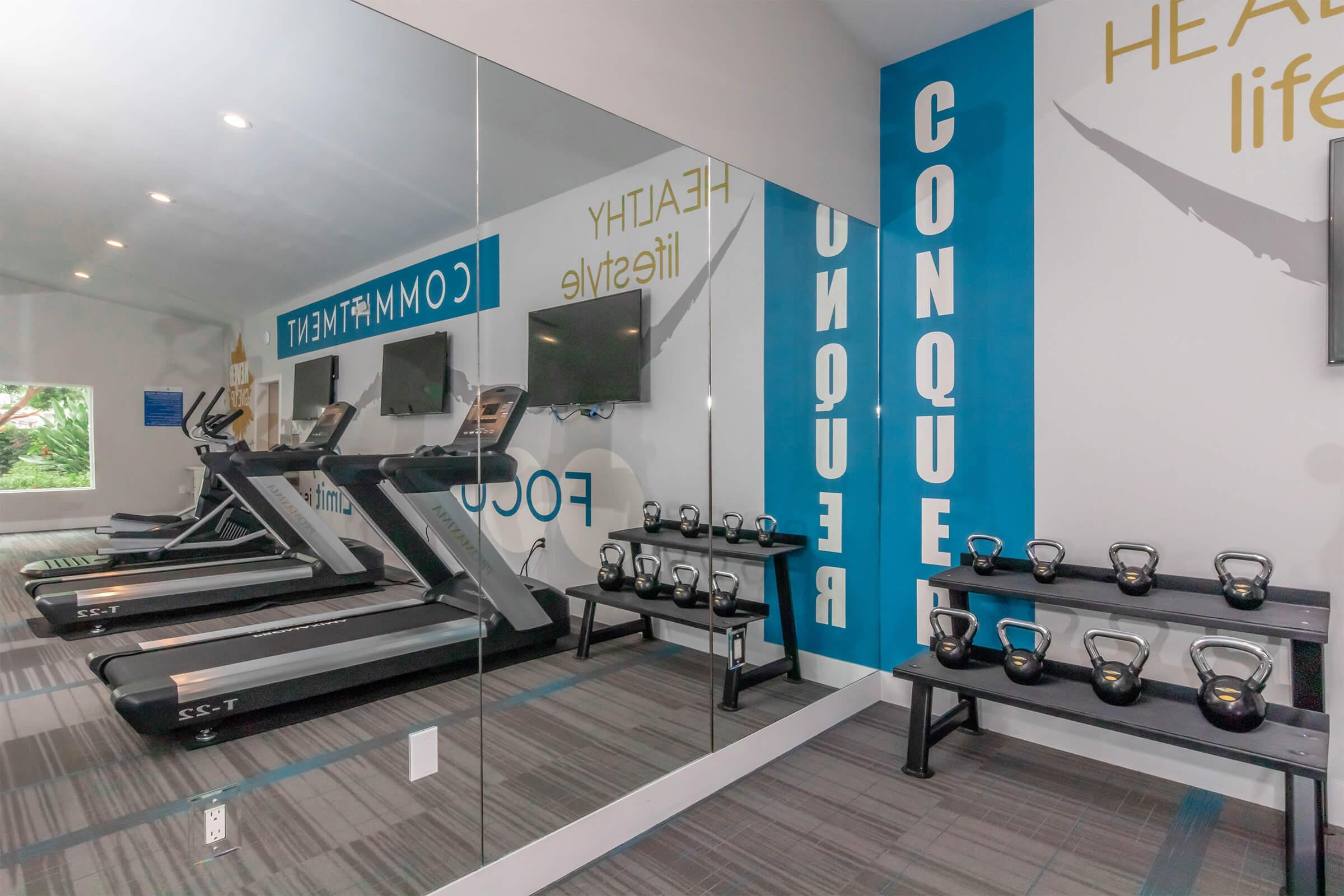 A modern fitness room featuring treadmills and a wall-mounted TV, with motivational text on the walls. A set of kettlebells is displayed on a rack, and large mirrors enhance the space, providing an open and inviting atmosphere for exercise.
