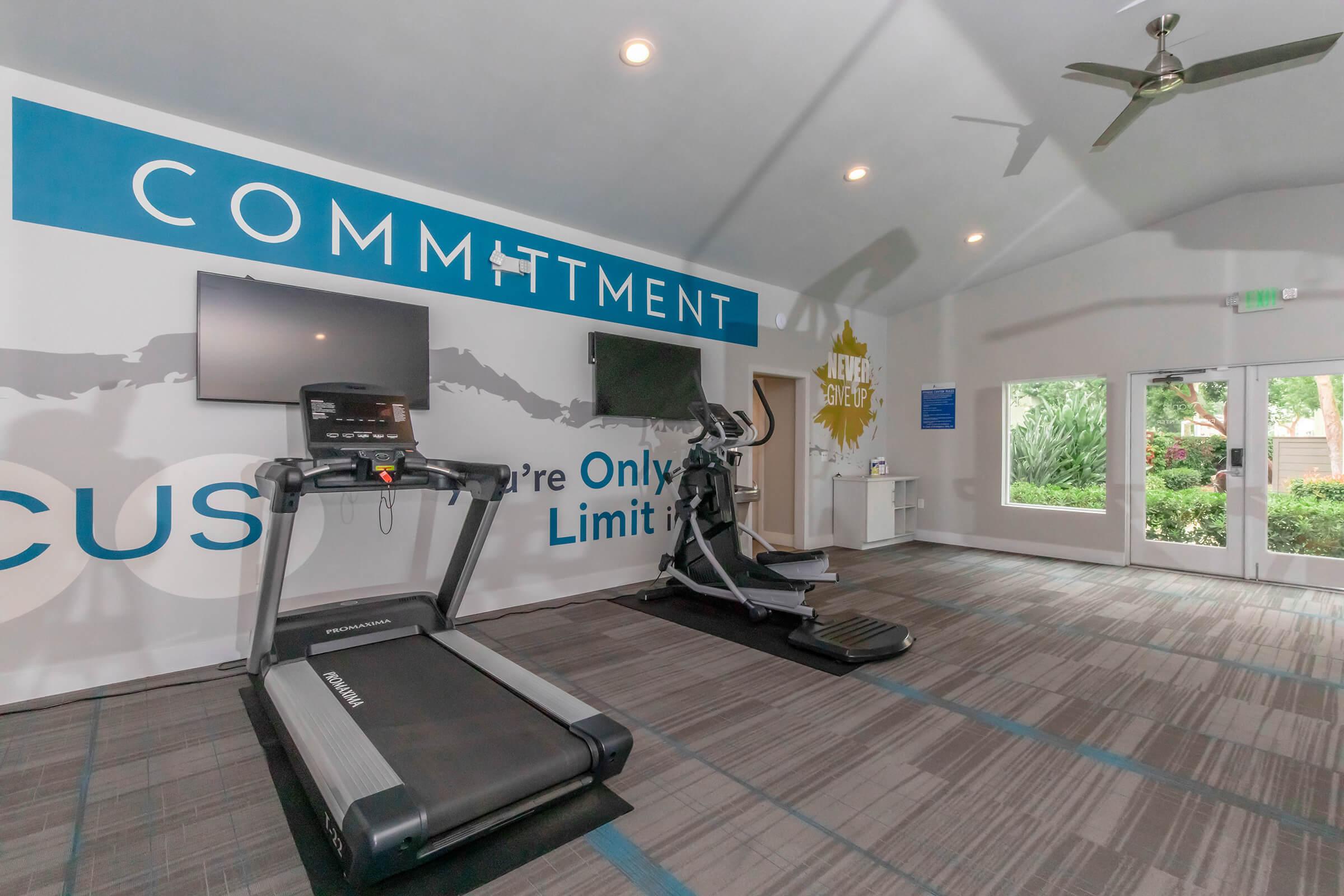A modern gym interior featuring a treadmill and an elliptical machine, with motivational text on the wall stating "Commitment" and "You're Only Limit Is." There are two mounted TVs on the wall, and large windows allowing natural light to enter, framed by lush greenery outside. The flooring is carpeted in a blue and gray pattern.