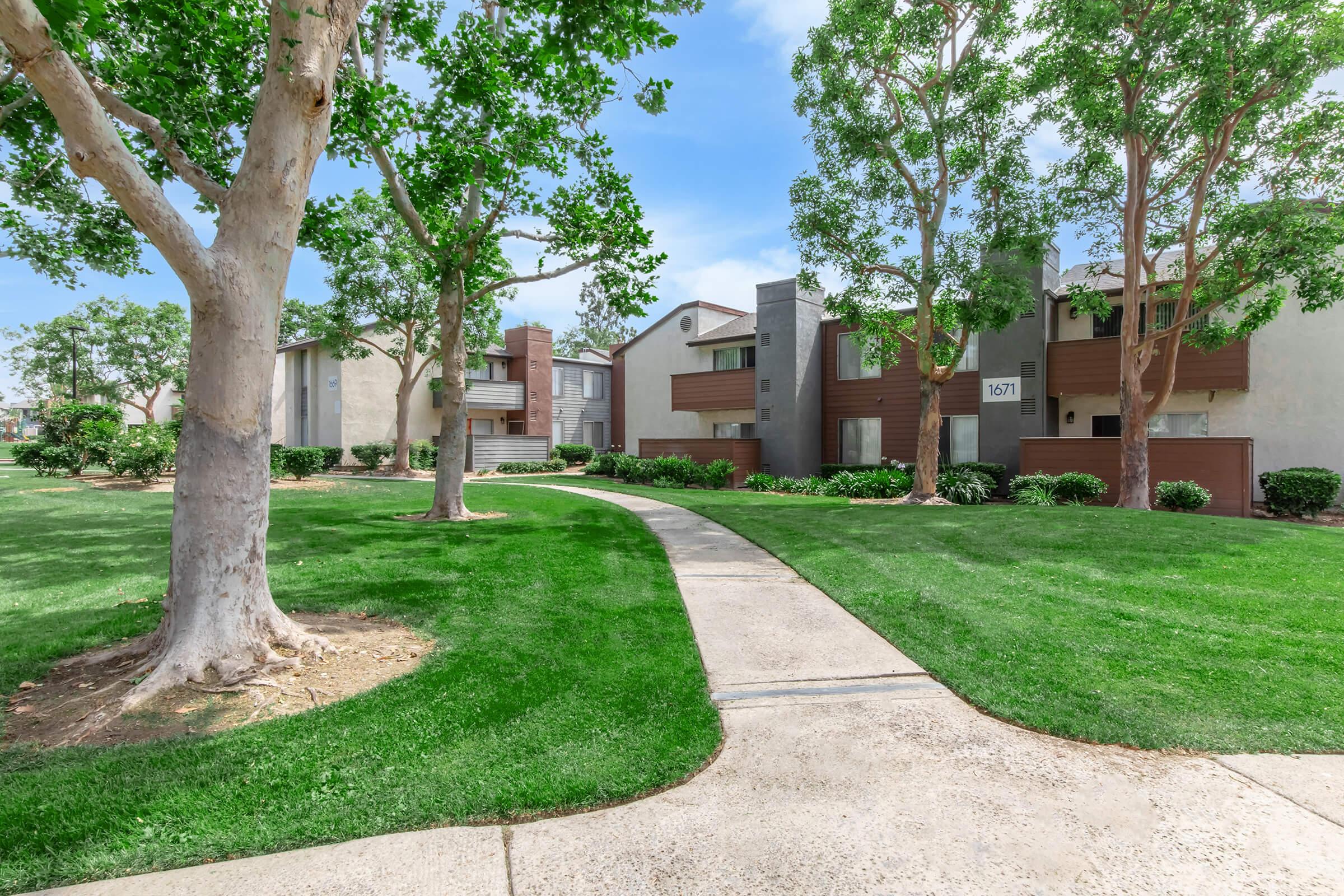 A pathway leads through a well-maintained green lawn lined with trees, leading to modern residential buildings. The buildings feature a mix of beige and gray exteriors with wooden accents. Bright blue sky and fluffy clouds create a serene atmosphere in the residential area.