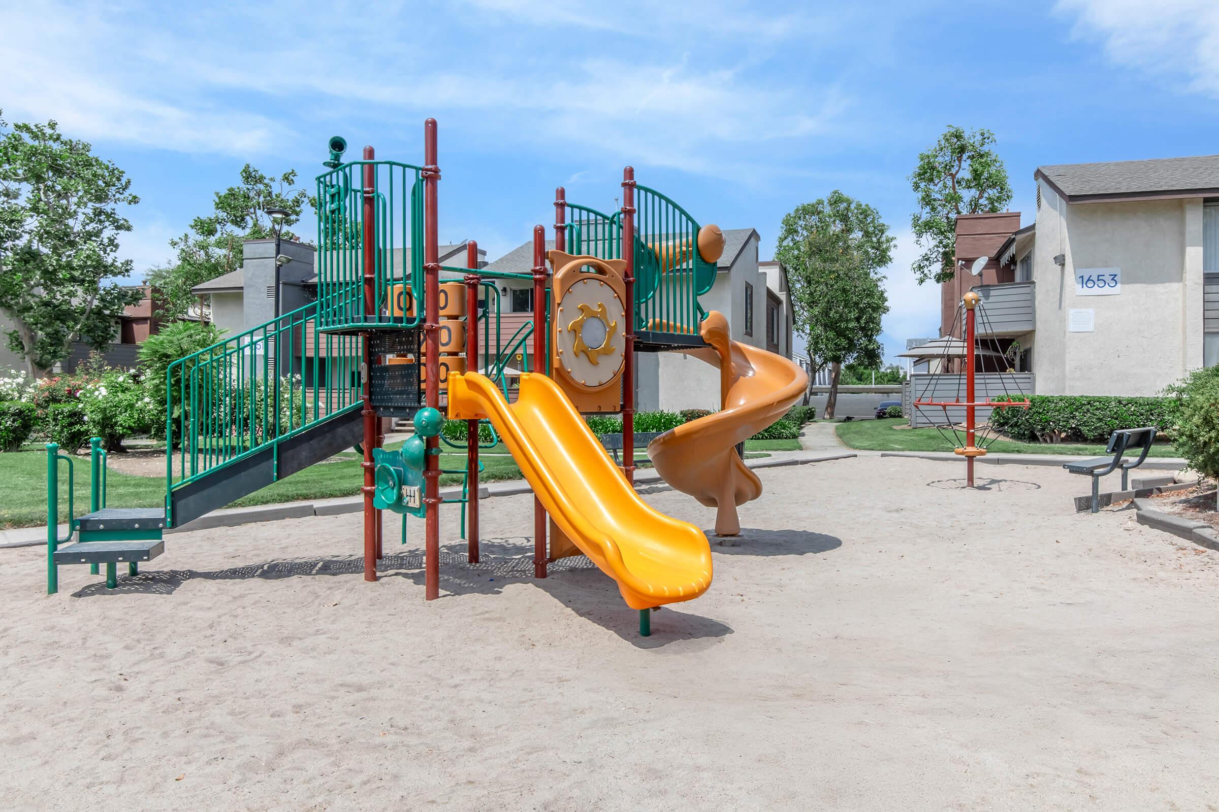 Colorful playground equipment featuring a green and orange slide, climbing structures, and a sandbox area. Surrounding grassy areas with benches and residential buildings in the background under a blue sky.