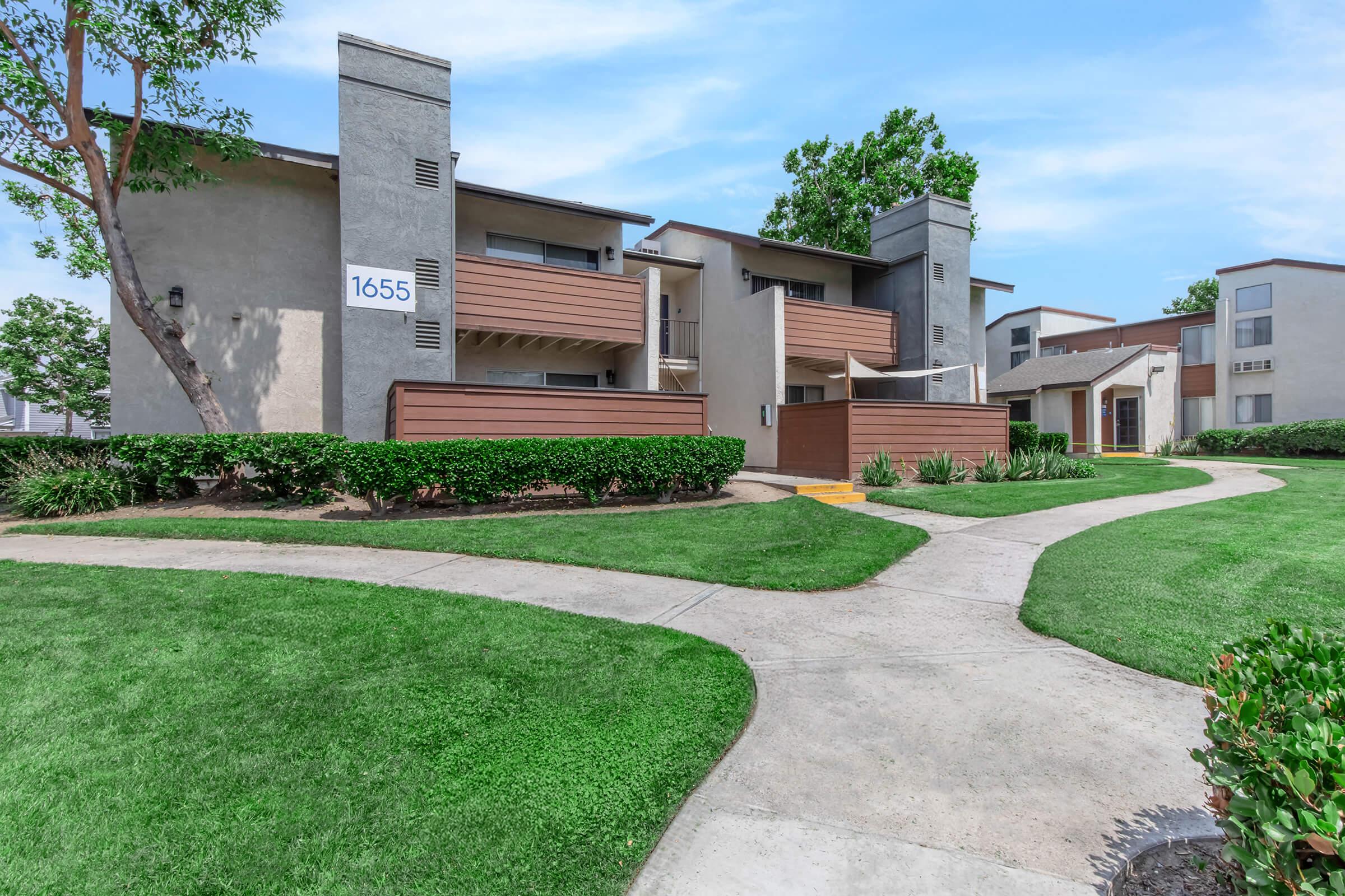 Exterior view of a residential apartment complex featuring modern architecture, with two-story buildings, landscaped lawns, and walkways. The image shows a building with the number "1655" prominently displayed and greenery surrounding the pathway.