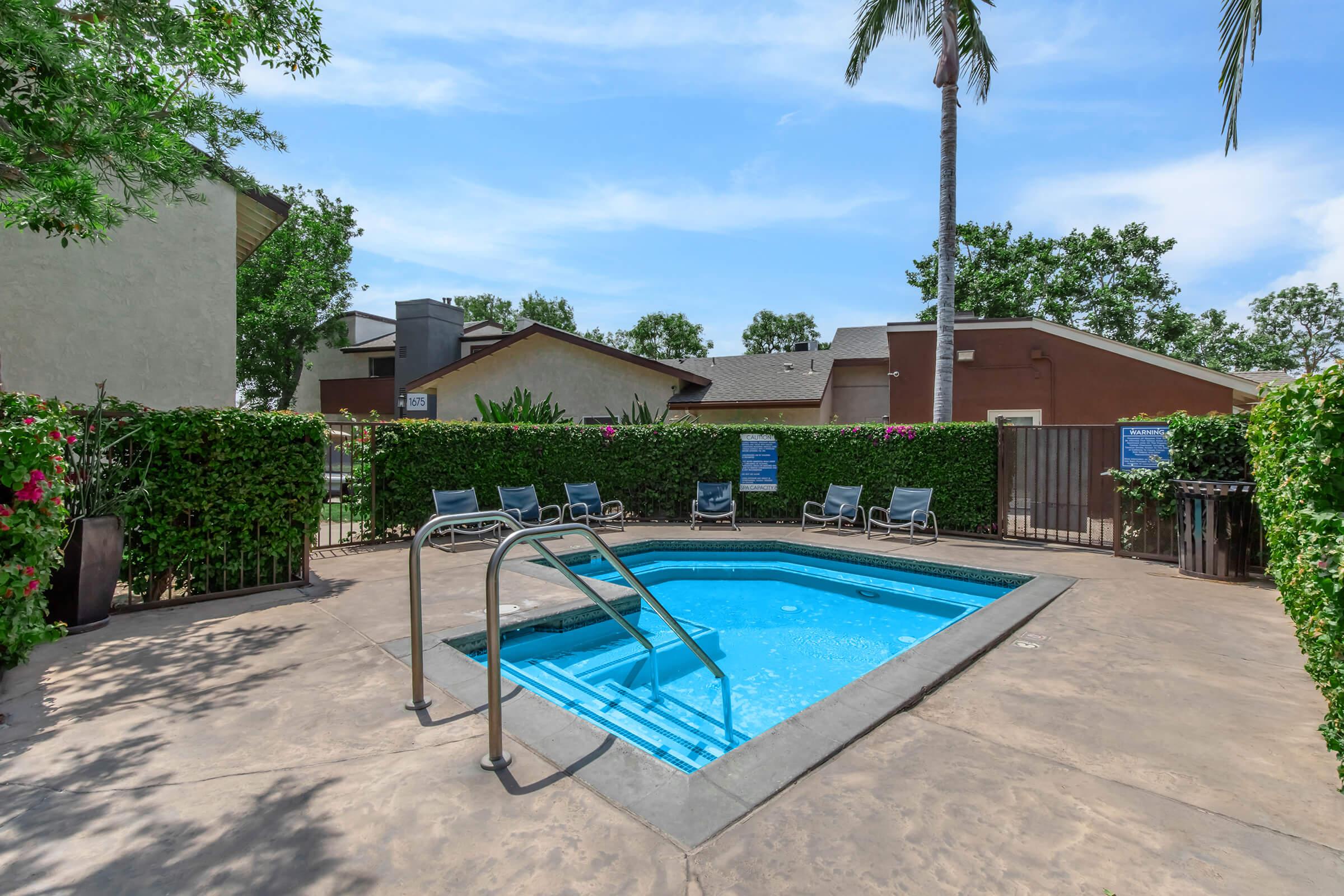 A small, inviting pool surrounded by greenery and lounge chairs, located in a sunny outdoor space. There are palm trees nearby, and a gated area with a fence in the background, creating a relaxing atmosphere. The sky is clear and blue, enhancing the tranquil setting.