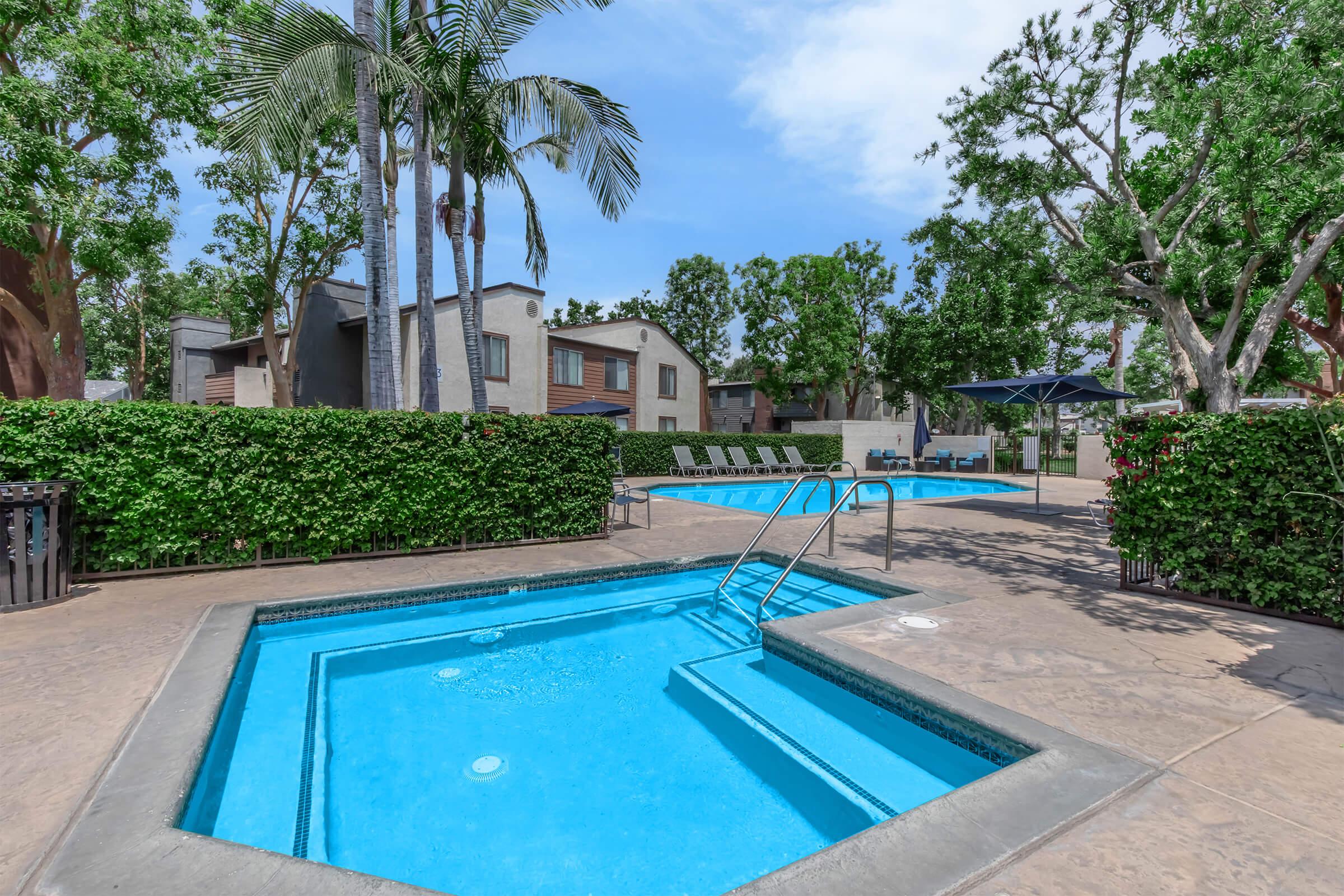 A vibrant outdoor area featuring multiple swimming pools surrounded by lush greenery and palm trees. One pool has a jacuzzi, while others are designed for leisure. There are lounge chairs and shaded areas, creating a relaxing atmosphere in a residential community.