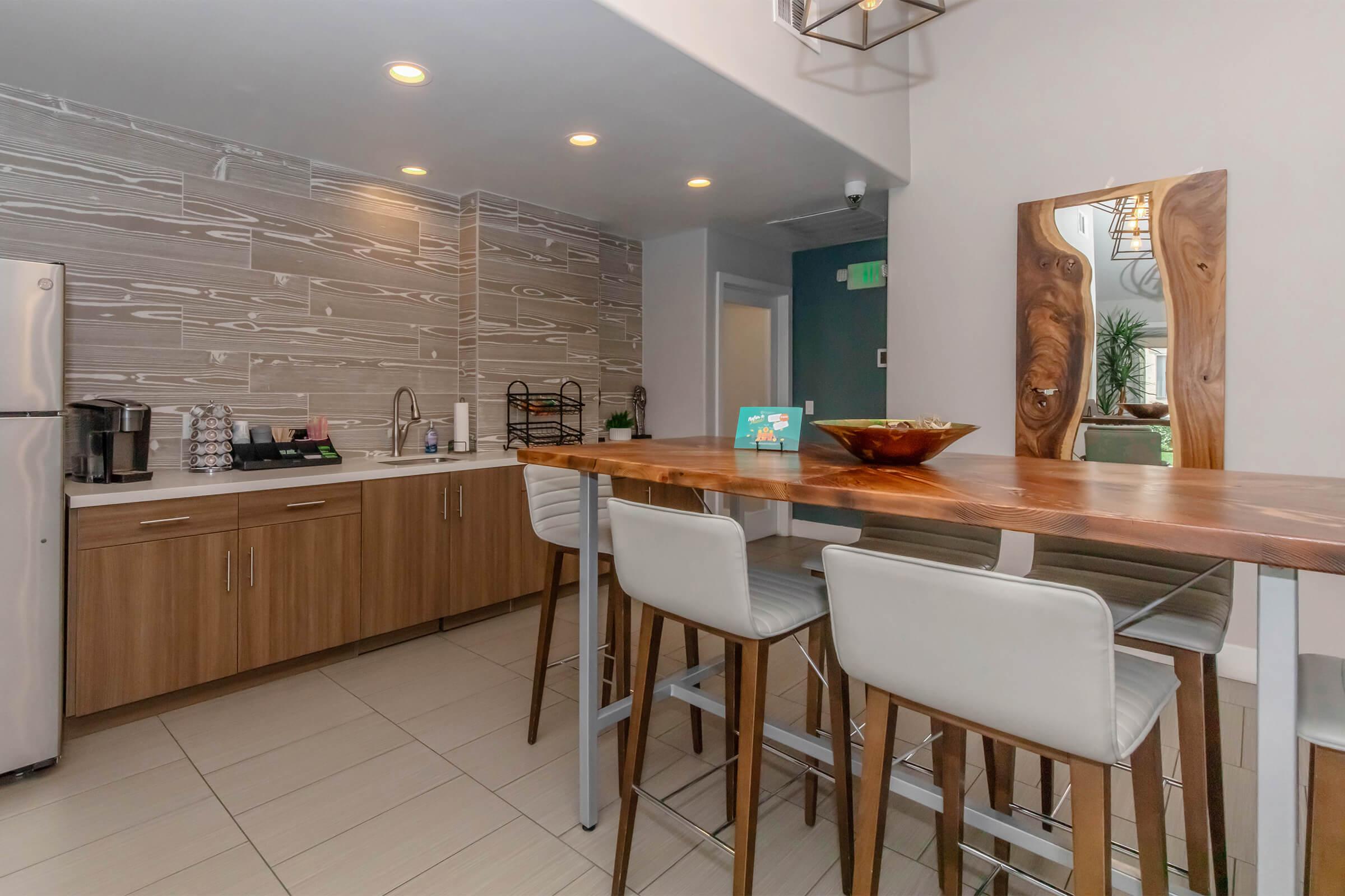 Modern kitchen with wooden cabinetry and a sleek countertop. Includes a silver refrigerator, coffee maker, and sink. A wooden dining table with white bar stools is in the foreground, and the wall features a textured wood pattern. Soft overhead lighting creates a warm ambiance. A small plant is visible in the background.