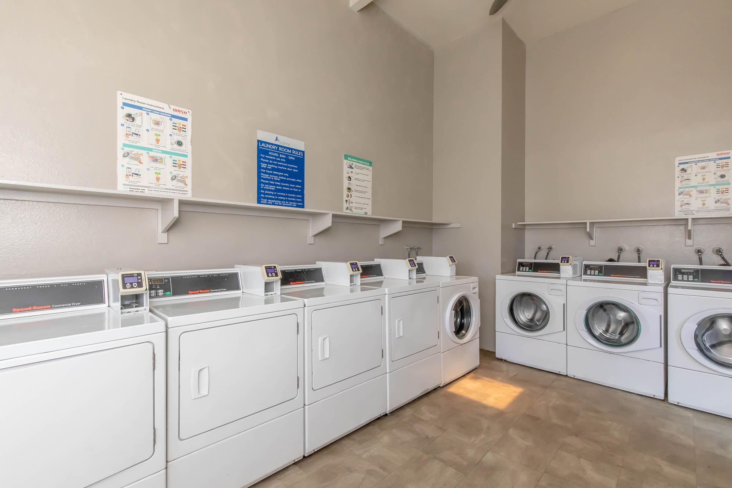 A clean and well-organized laundromat with multiple white washers and dryers arranged along the walls. Informative posters are displayed on the walls, providing laundry instructions and guidelines. Natural light enters the room, enhancing the bright and tidy atmosphere.