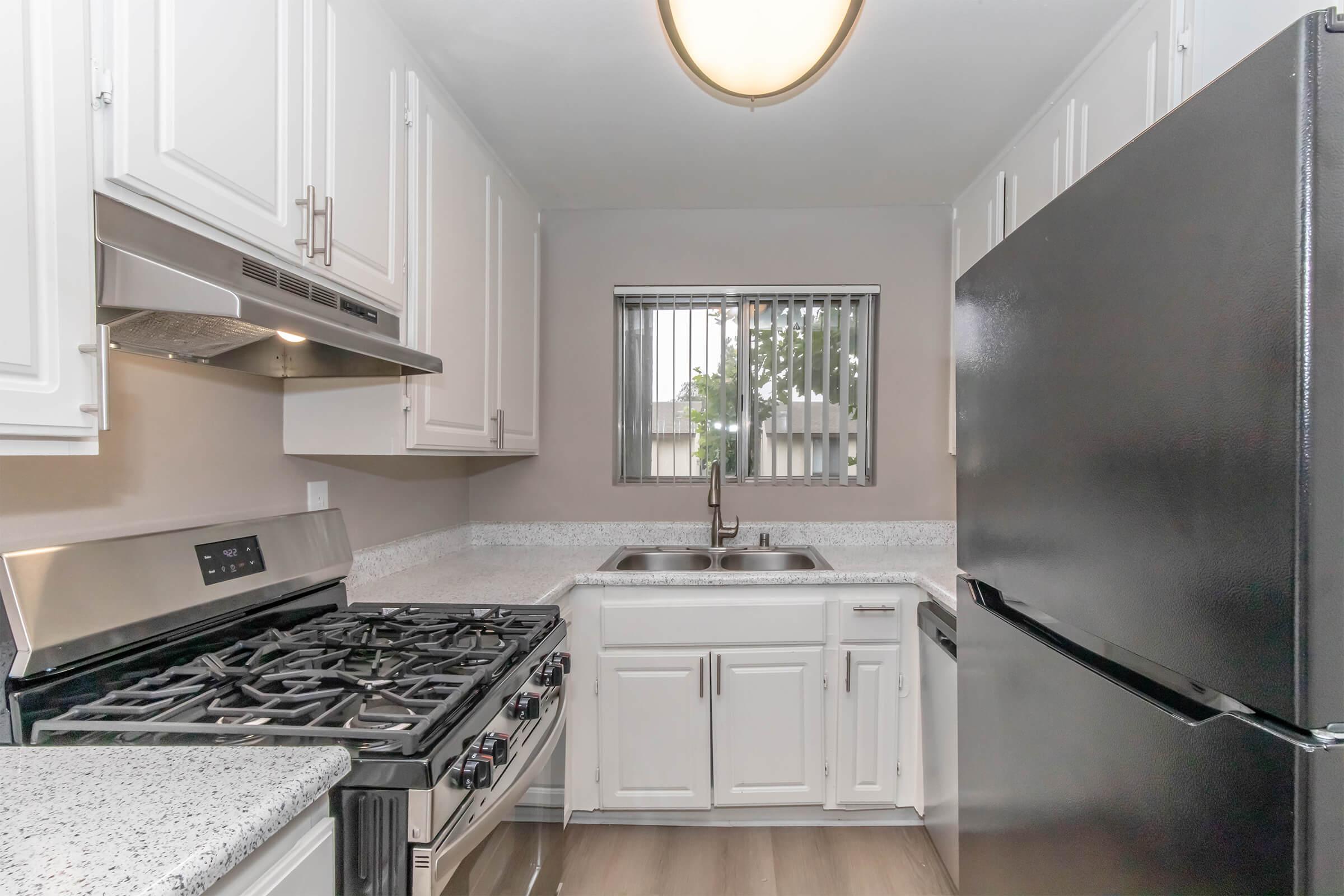 Modern kitchen with white cabinetry, stainless steel appliances including a gas stove and a range hood, a black refrigerator, and a double sink beneath a window. The countertop features a light-colored granite finish, and the flooring is light wood.