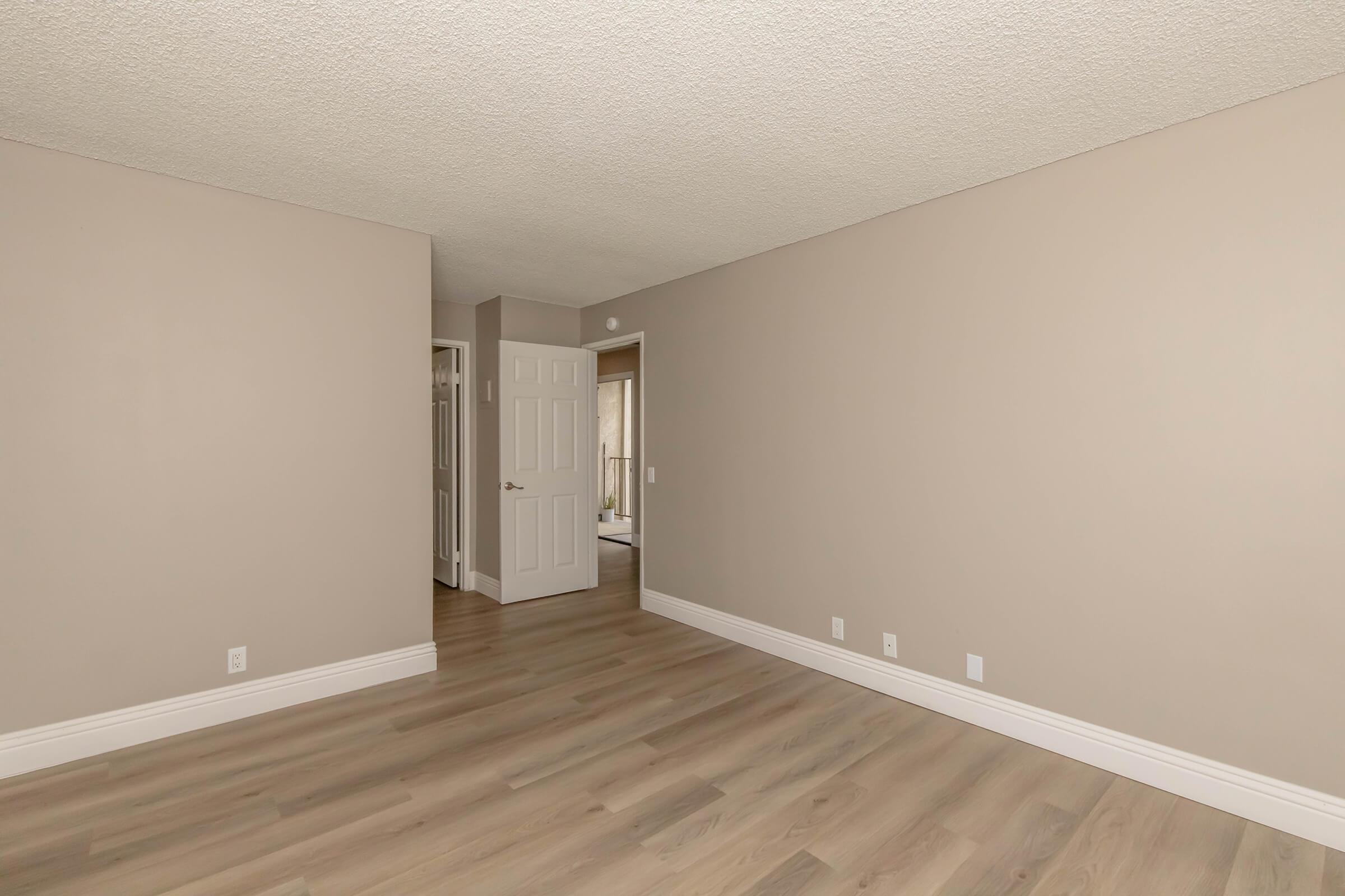 A spacious, empty room with light beige walls and new laminate flooring. The room features a doorway leading to another area, natural light coming through an unseen window, and a seamless design with no furniture, creating a clean and modern aesthetic.