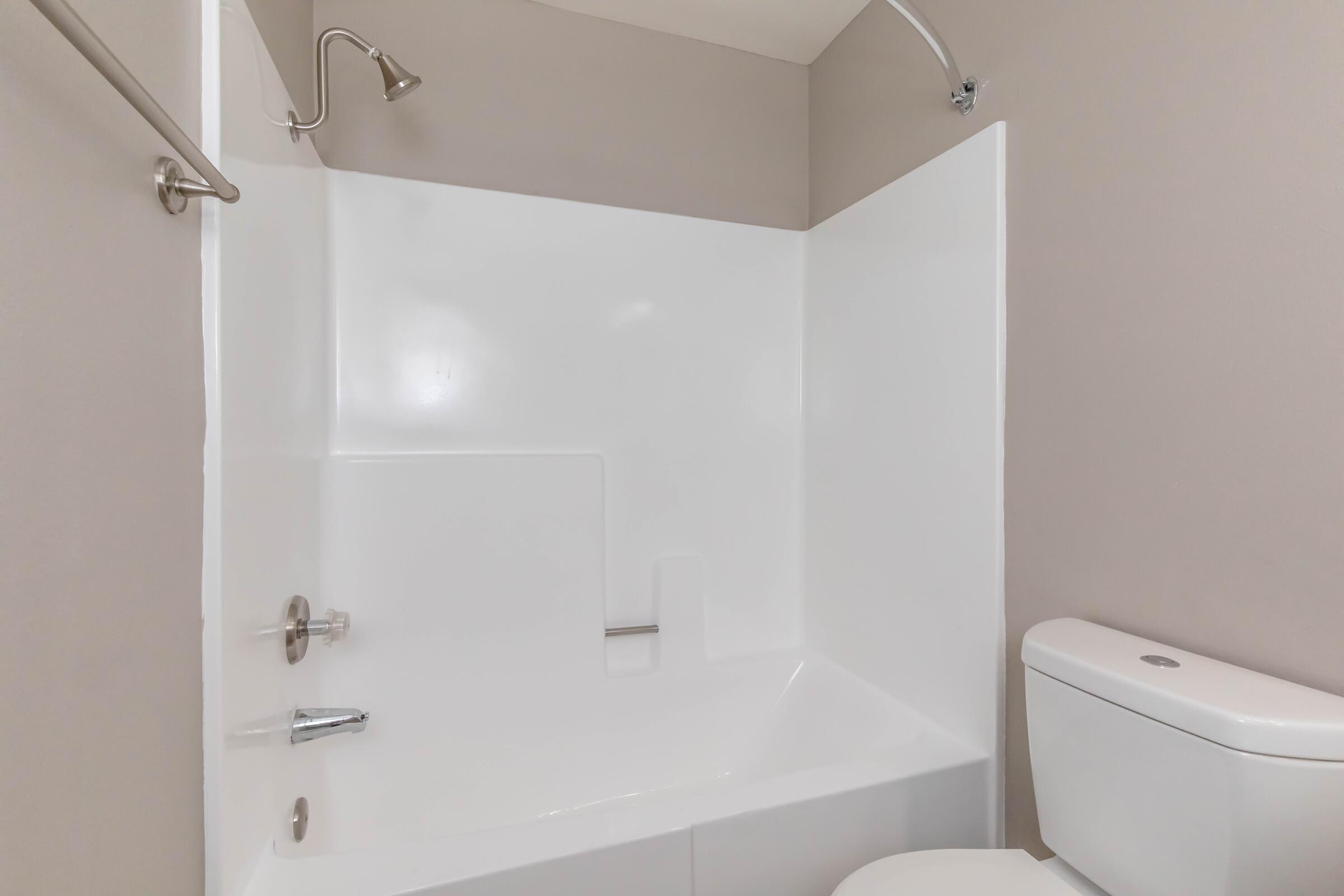A clean, modern bathroom featuring a white bathtub and shower, a chrome faucet, and a toilet. The walls are painted in a neutral beige color, creating a simple and functional space.