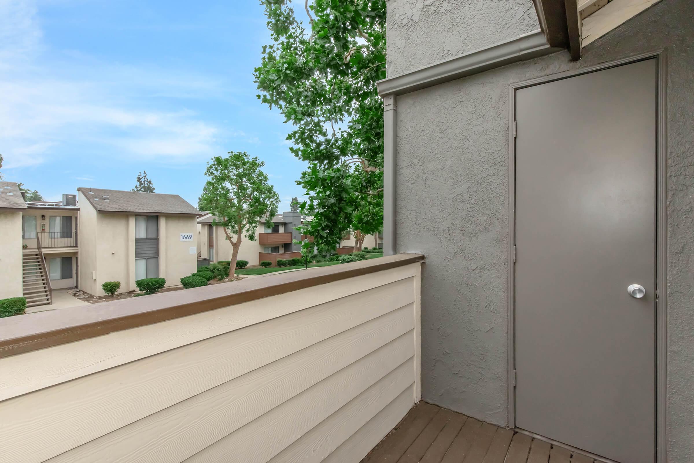 A view from a balcony overlooking a residential area. There are several buildings with beige exteriors and green landscaping. A gray door is visible on the right side, and trees are scattered throughout the neighborhood under a clear blue sky.