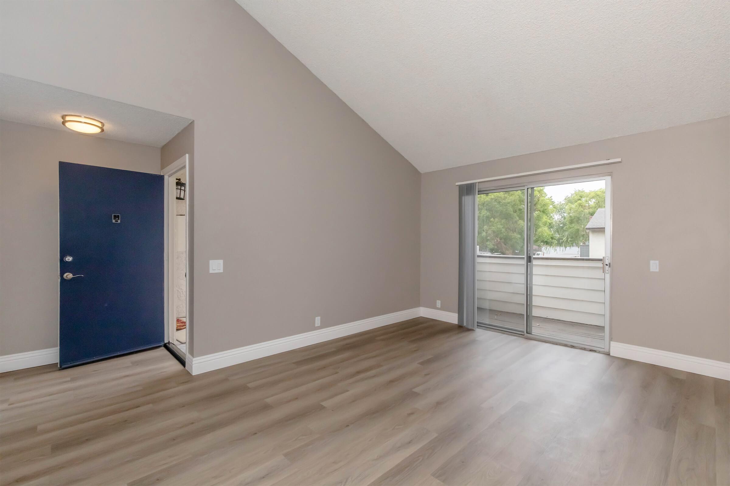 A spacious, unfurnished living room with high ceilings and large windows. The room features light wood flooring and neutral-colored walls. A sliding glass door leads to a small outdoor area, while a dark blue door is visible near the entrance.