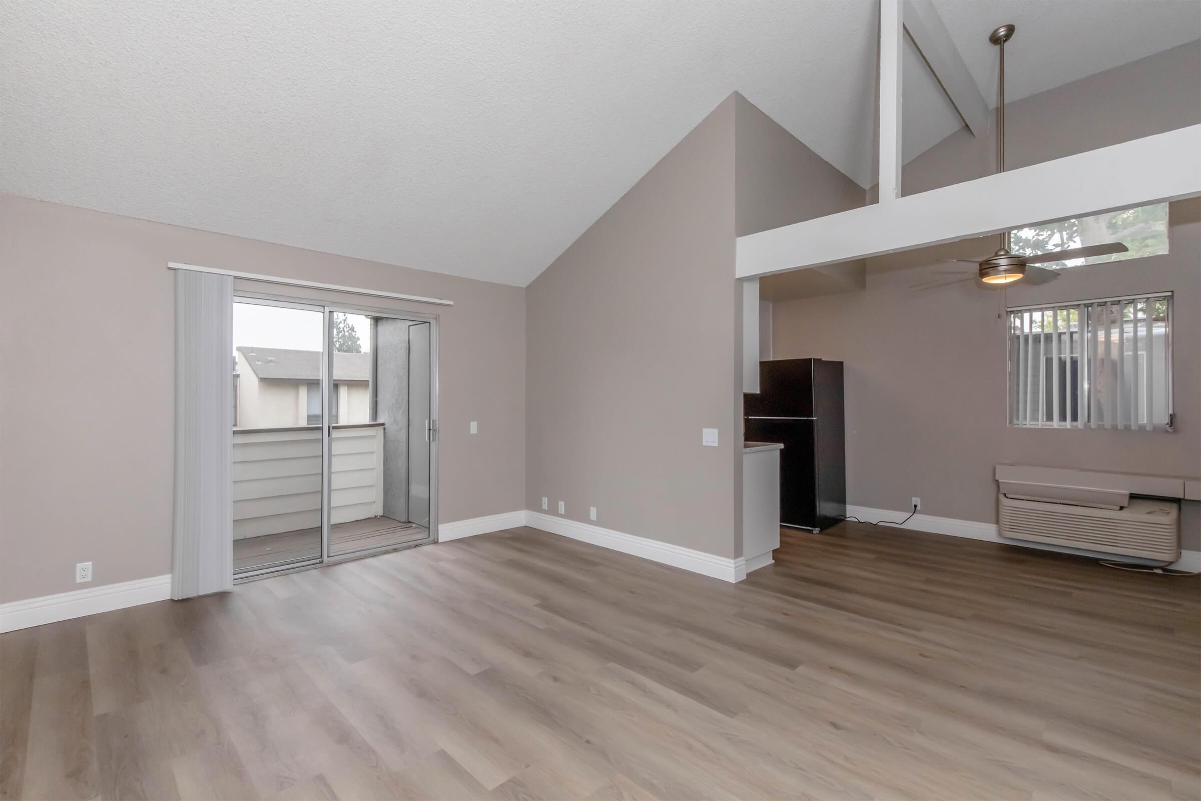 Spacious interior of a living area featuring light gray walls and wooden flooring. A large sliding glass door leads to a balcony. On the right, a ceiling fan is visible, along with a black fridge and an air conditioning unit. The vaulted ceiling adds height and openness to the room.