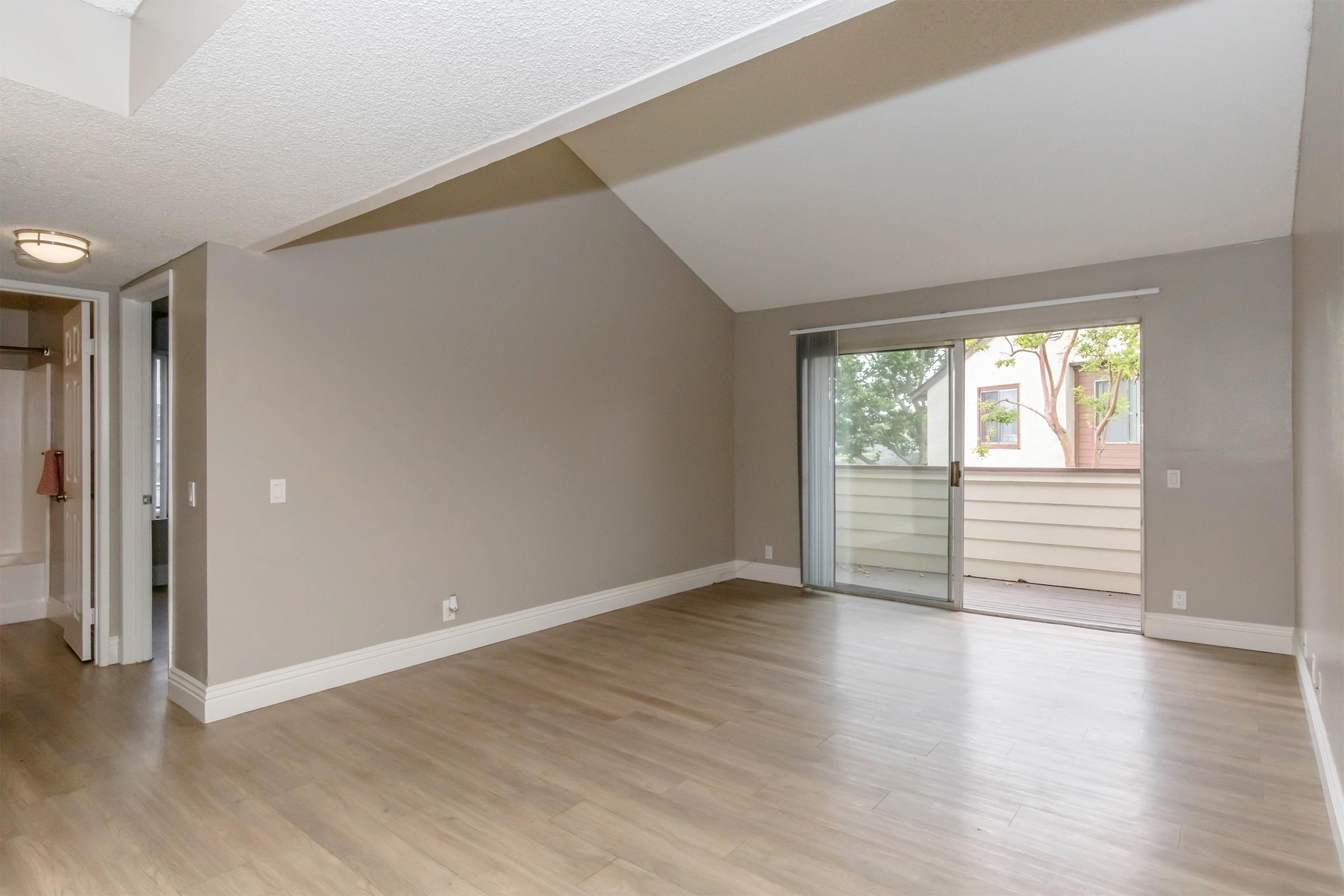 A spacious, empty living room featuring light-colored walls and hardwood flooring. A large sliding glass door leads to a balcony with views of trees outside. The room has a vaulted ceiling and includes an open layout with natural light. A doorway on the left leads to a bathroom area.
