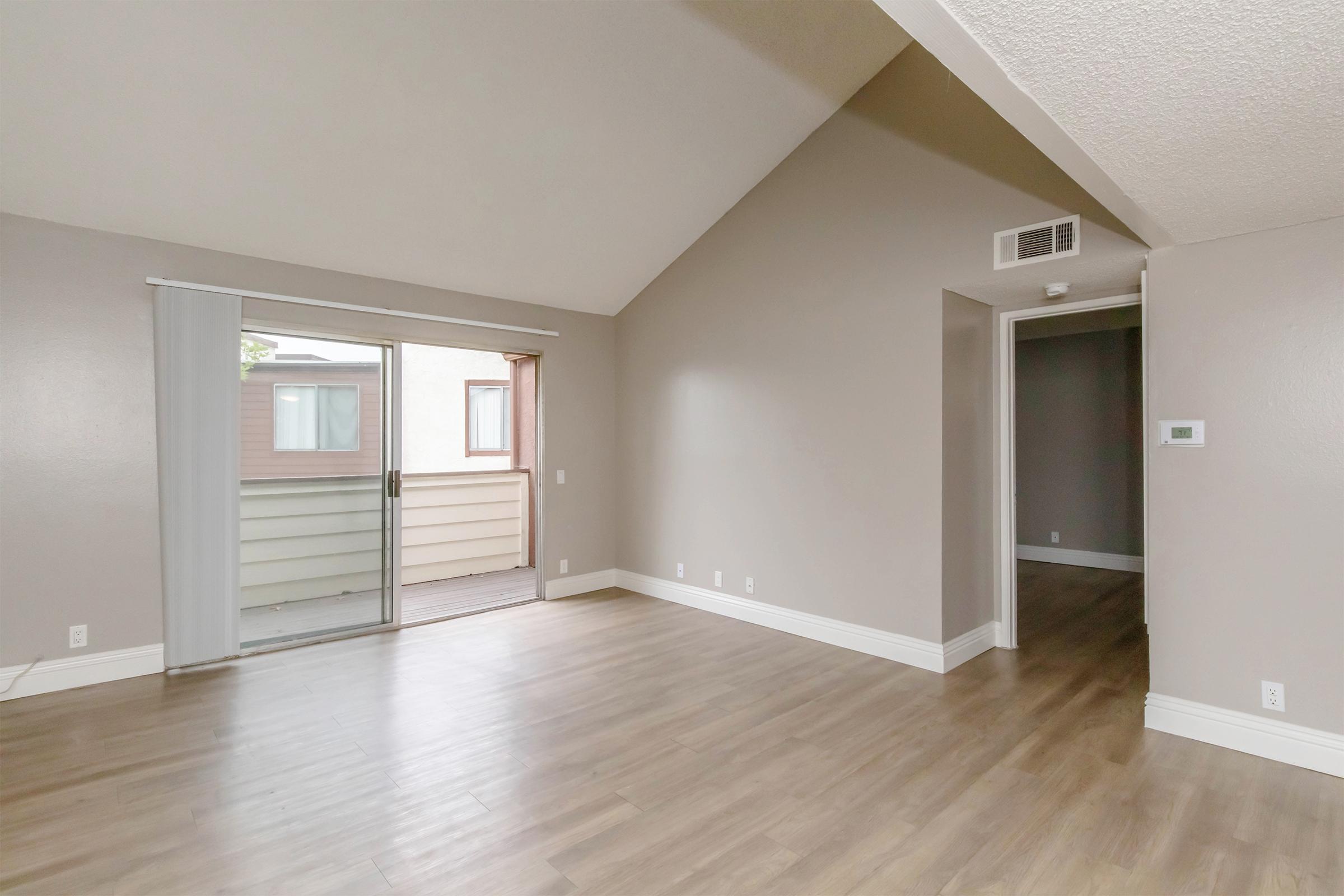 Spacious living area featuring light-colored walls and a vaulted ceiling. A large sliding door leads to a balcony, allowing natural light to fill the room. The flooring is light wood, and there's a hallway to the right leading to other areas of the home.
