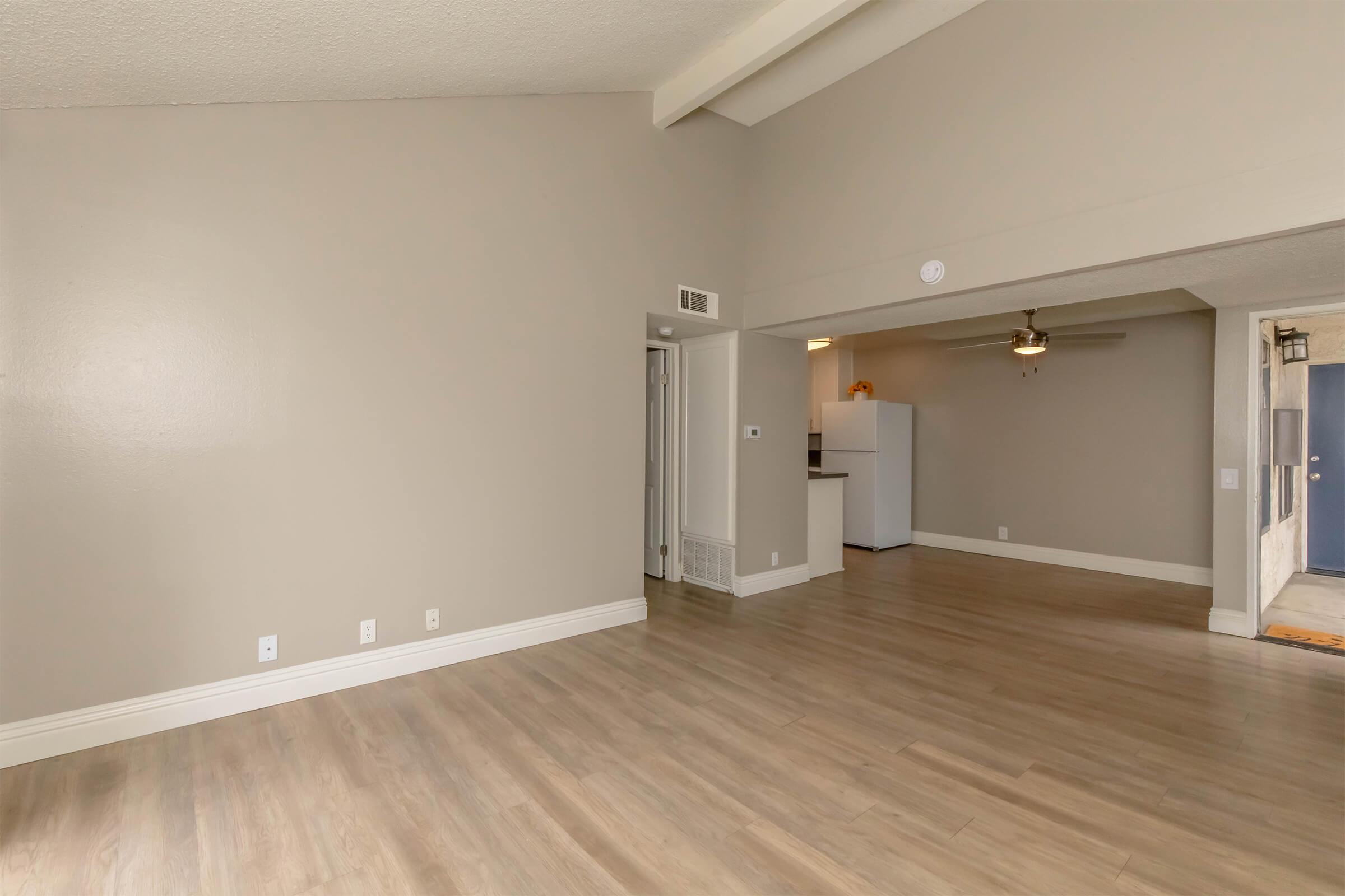 Spacious living area with light beige walls and hardwood-style flooring. The room features a high ceiling and a ceiling fan. A doorway leads to a kitchen area in the background, which includes white appliances. The overall ambiance is bright and inviting, with natural light streaming in from unseen windows.