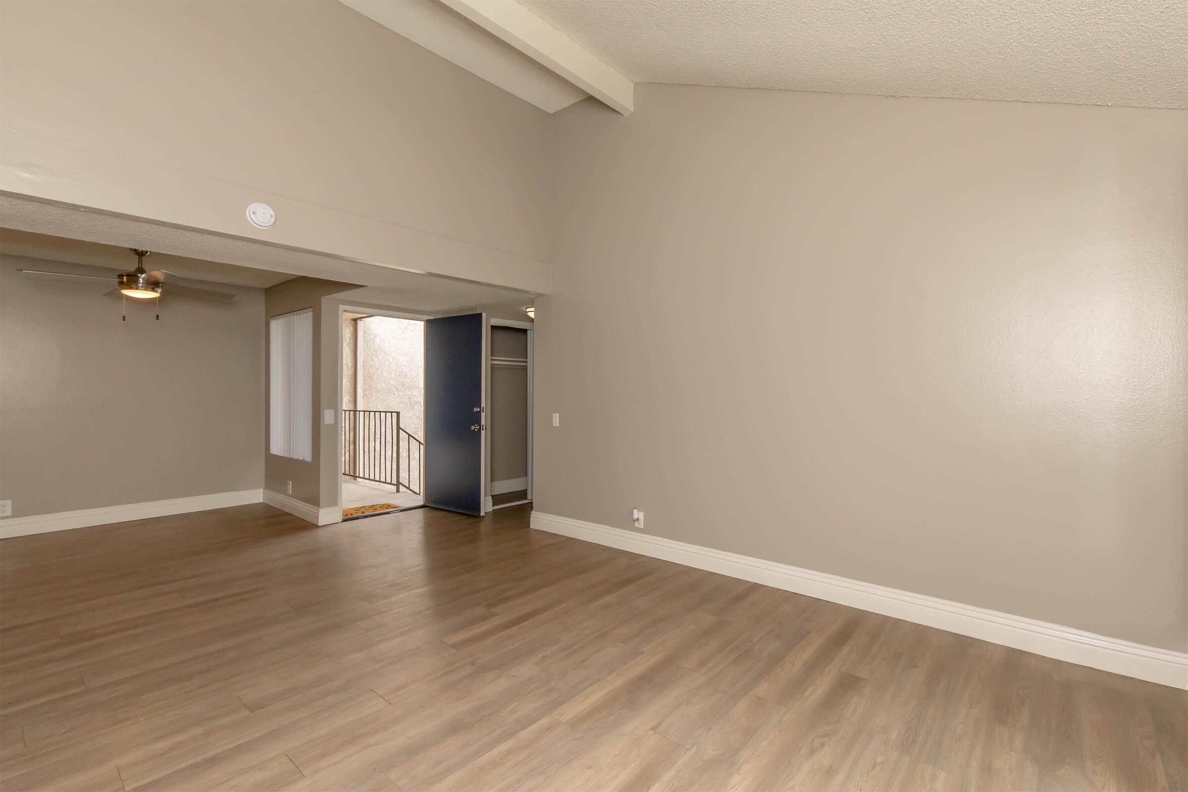 A spacious, well-lit living area with light-colored walls and wooden flooring. The room features a ceiling fan, and there's an opening leading to a hallway with a blue door. Natural light enters from the hallway, creating an inviting atmosphere.