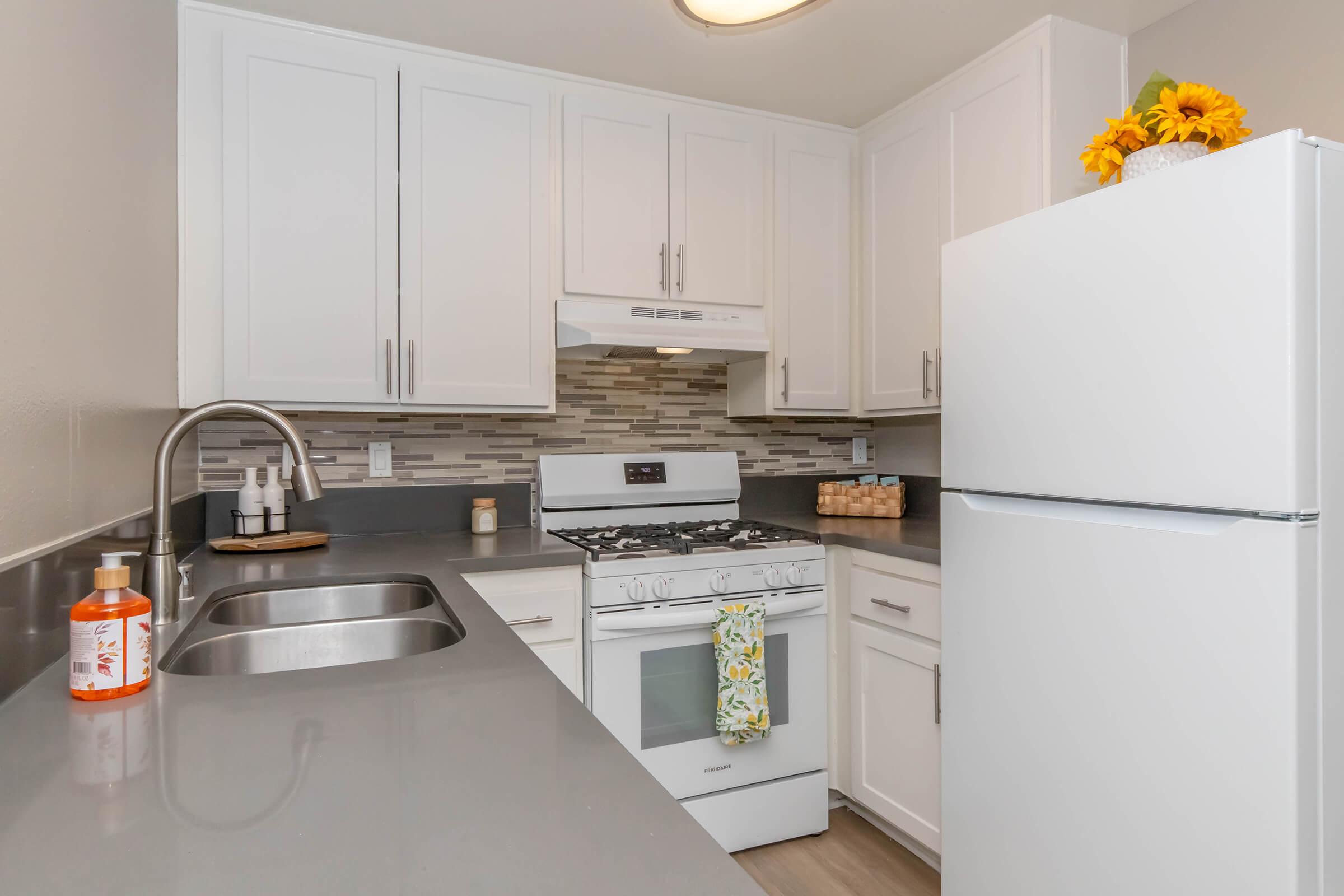 Modern kitchen featuring white cabinetry, a stainless steel sink with a sleek faucet, a gas stove under a vent hood, and a refrigerator. The countertop is gray with a textured design. Decorative items include a bottle of oil and a small vase with sunflowers. A tile backsplash adds a stylish touch.