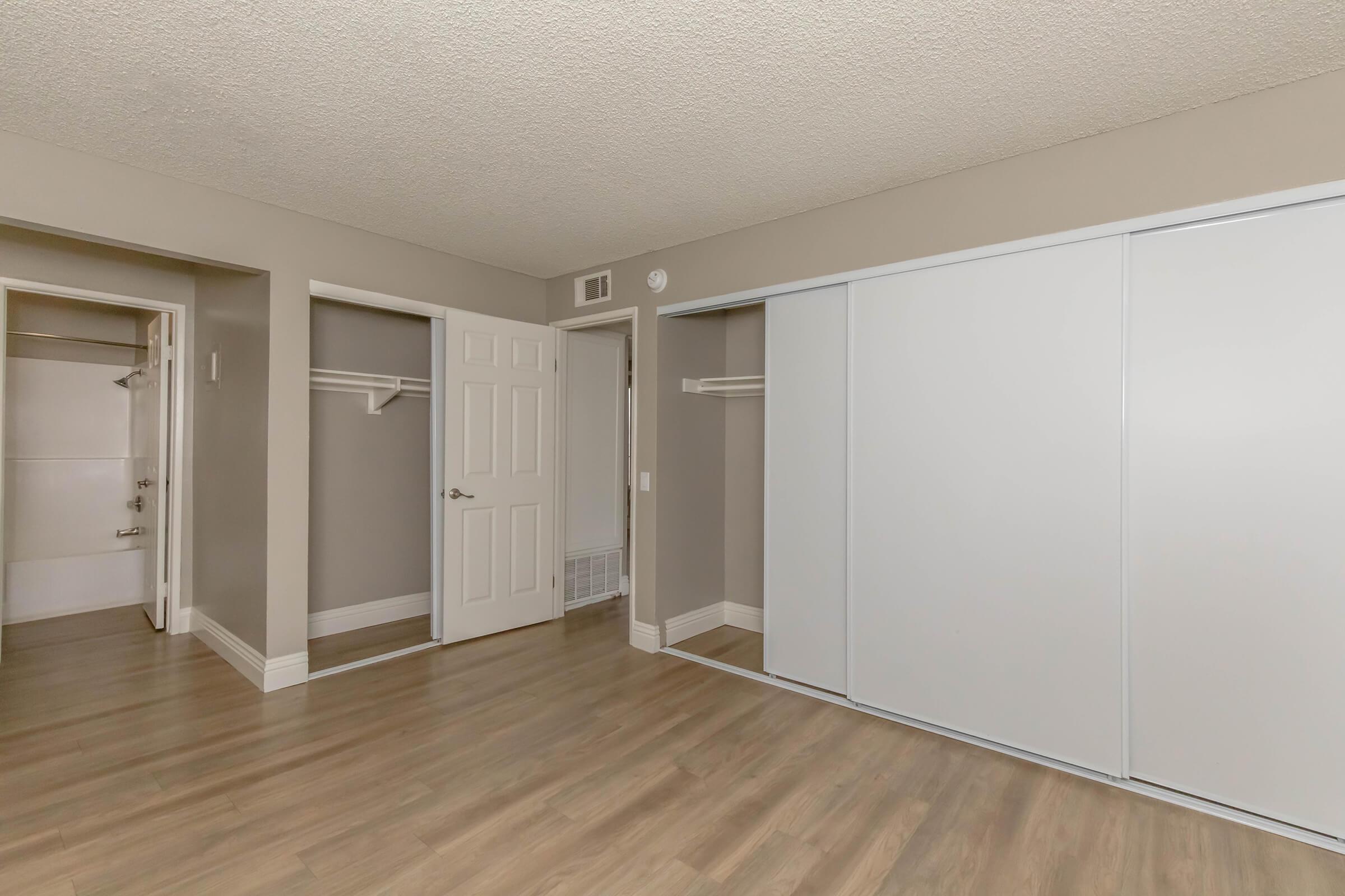A spacious, empty room featuring beige walls and wood laminate flooring. There are two closet spaces with sliding doors and a door leading to another area. The lighting is neutral, and the overall atmosphere is clean and minimalistic, suitable for various furniture arrangements.