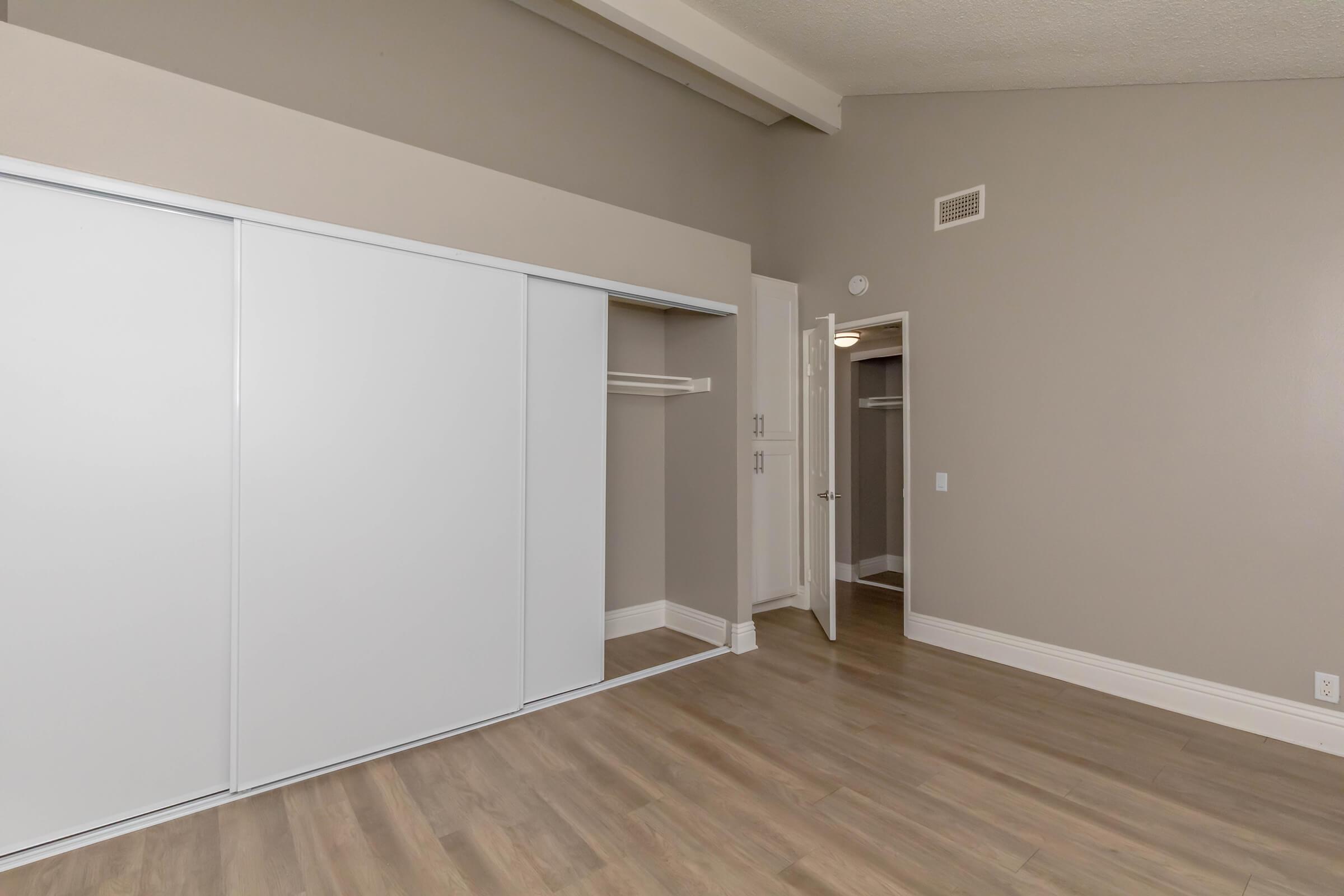 A modern, empty bedroom featuring a sliding door closet with white panels, light-colored walls, and wooden flooring. A separate door leads to another space, with ample natural light and a simple, contemporary design.