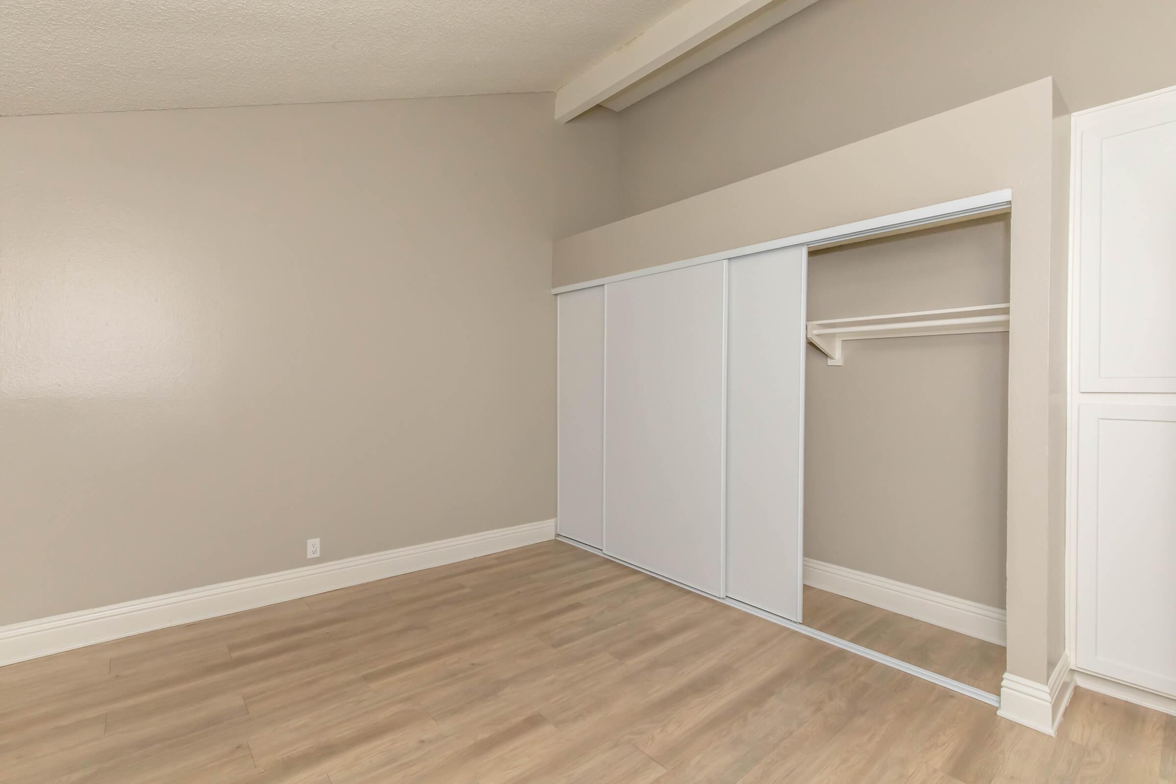 Empty room featuring light gray walls and a wooden floor. A sliding closet door is positioned on the left, revealing a simple shelving unit inside. The overall space is well-lit and minimalistic, creating an open and inviting atmosphere.