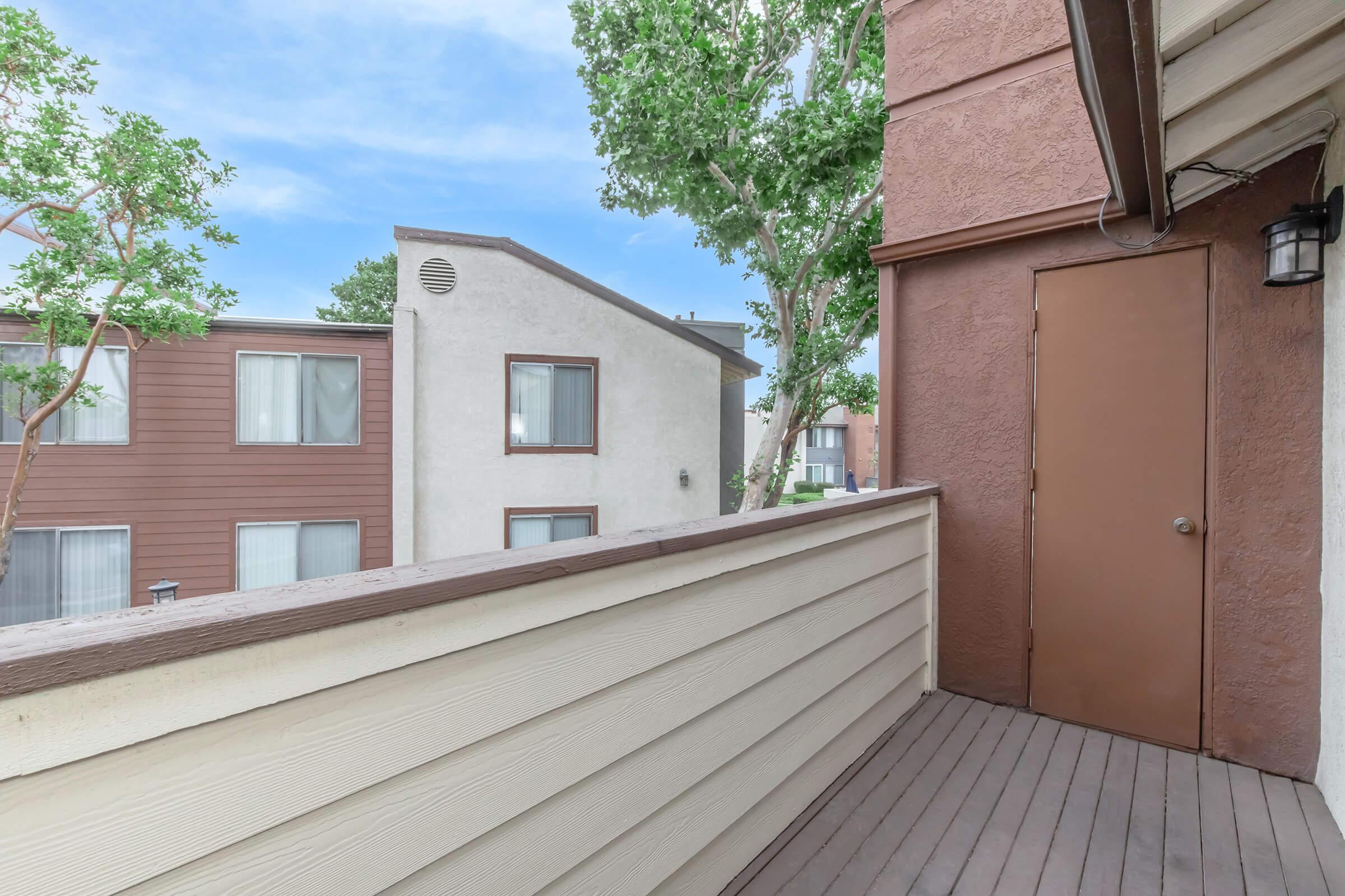 A balcony view with wooden flooring, featuring brown walls and a door on the left. In the background, there are two buildings with large windows and a green tree providing some shade. The sky is clear and blue, adding a bright, open feel to the outdoor space.