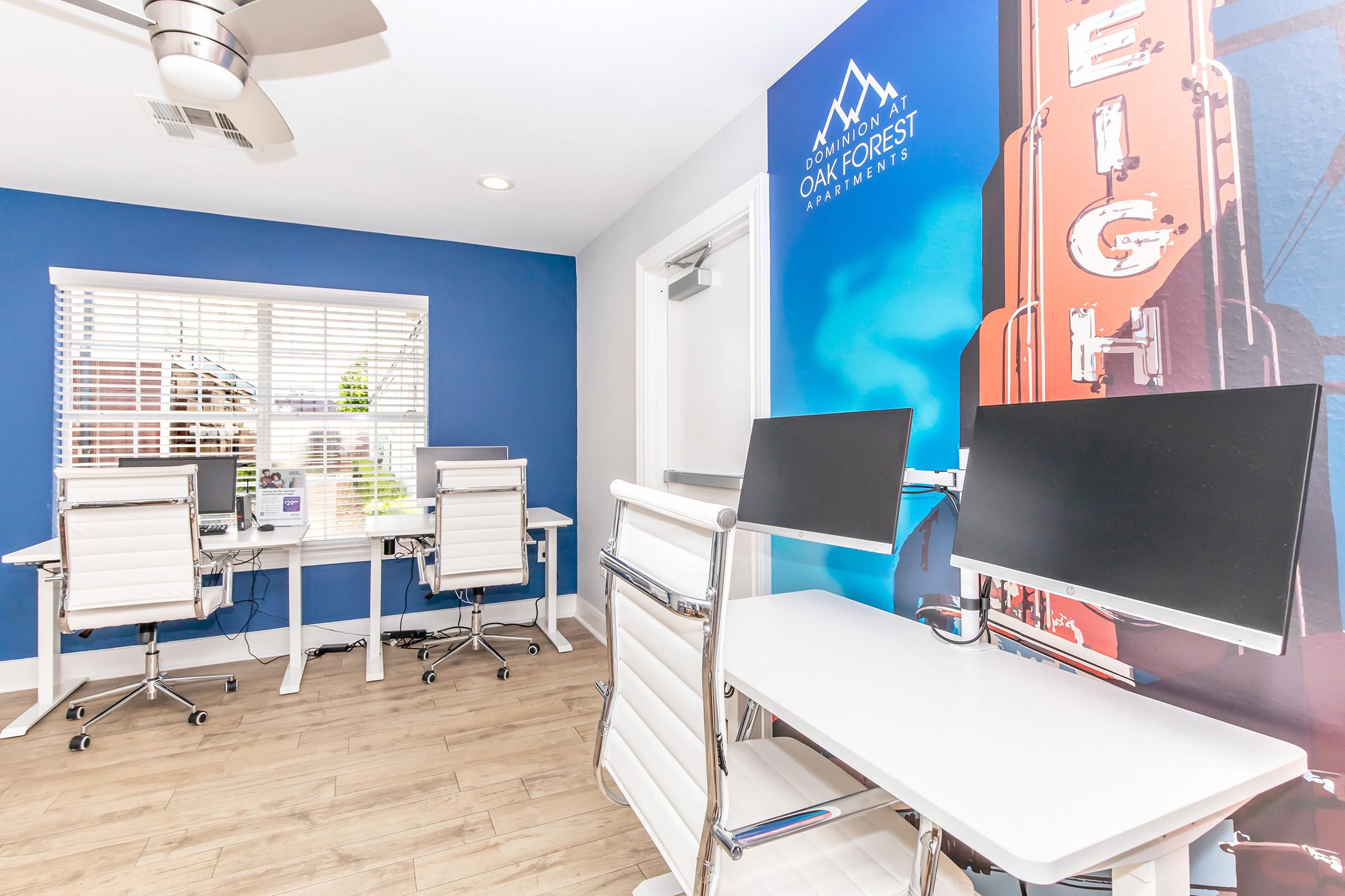 A modern office space featuring two white desks with computer monitors, silver chairs, and a blue accent wall. Large windows provide natural light, and a wall mural showcases a scenic design, enhancing the overall aesthetic. The floor is light-colored wood, creating a bright and inviting atmosphere.