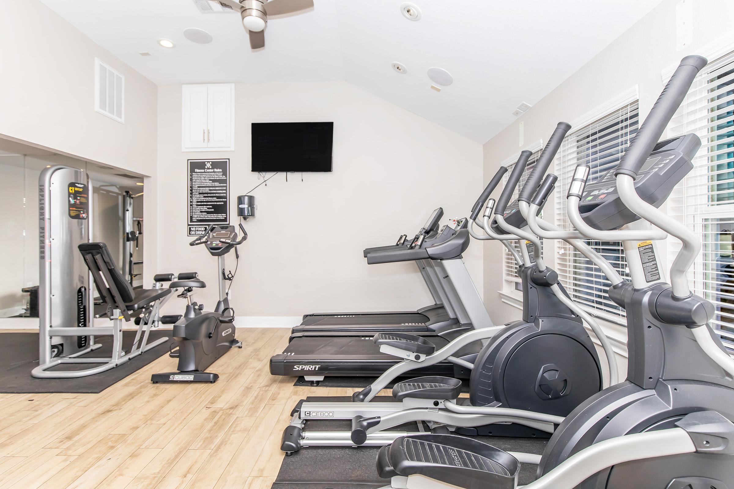 A well-lit gym featuring various exercise machines including treadmills, an elliptical, a stationary bike, and a multi-use weight machine. The room has large windows allowing natural light and a flat-screen TV mounted on the wall. The flooring is wooden with a dark rubber workout area.