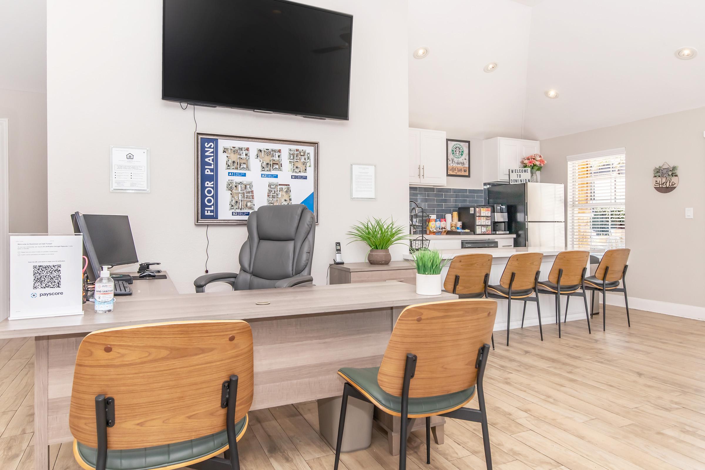 A modern office space featuring a reception area with a large TV on the wall, a desk with a chair, and several wooden chairs arranged in front. The desk has a plant and informational materials, while a kitchenette is visible in the background with cabinets and a coffee machine. Natural light fills the room.