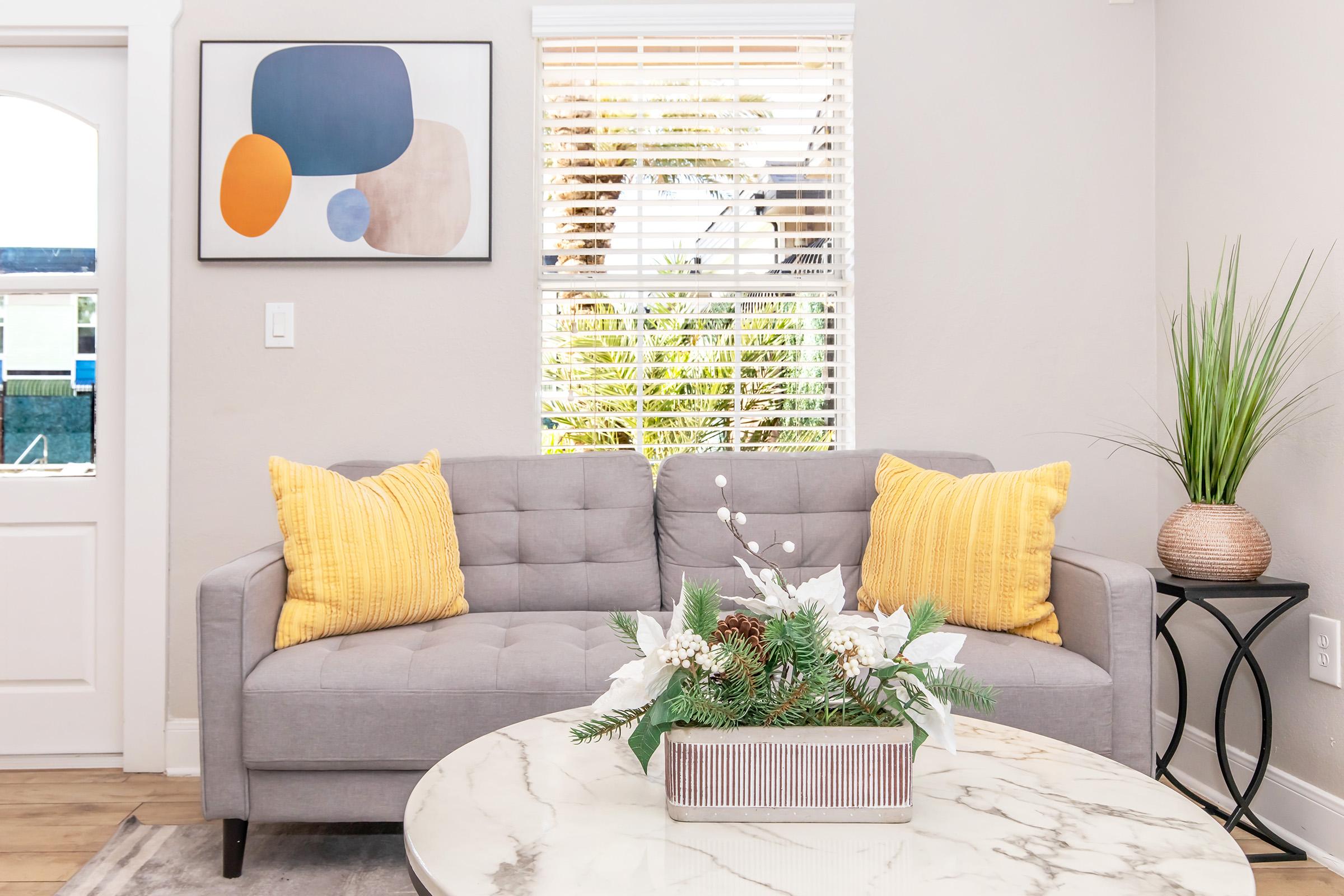A modern living room featuring a light gray sofa with two yellow cushions. In front of the sofa is a round marble coffee table with a decorative planter containing greenery and white flowers. A large window with blinds lets in natural light, and an abstract artwork hangs on the wall.