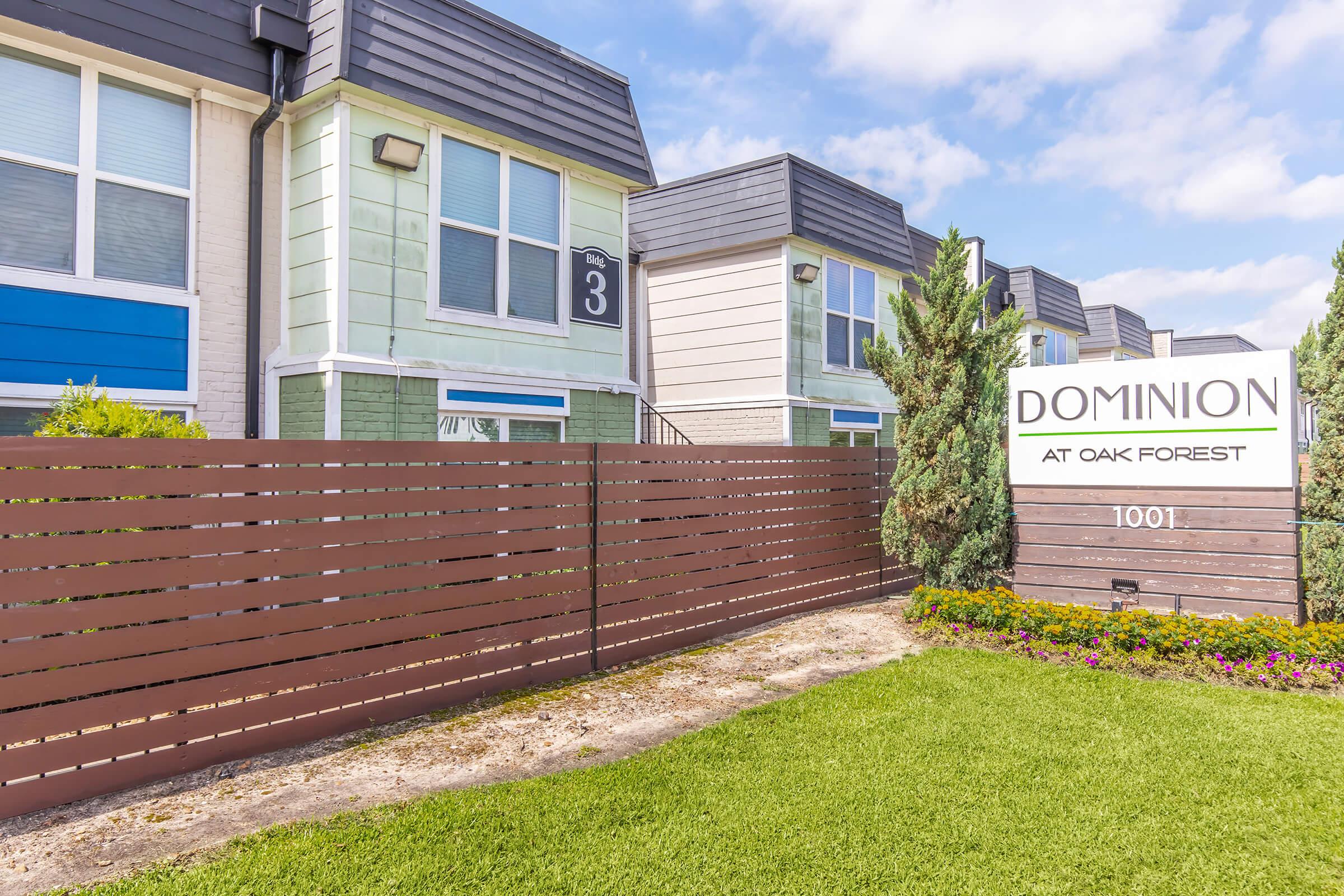 Exterior view of a multi-unit residential building complex named "Dominion at Oak Forest." The image shows two buildings with varying colors, a decorative wooden fence, and a sign with the address 1001. Green lawn and landscaping are visible in front of the buildings.