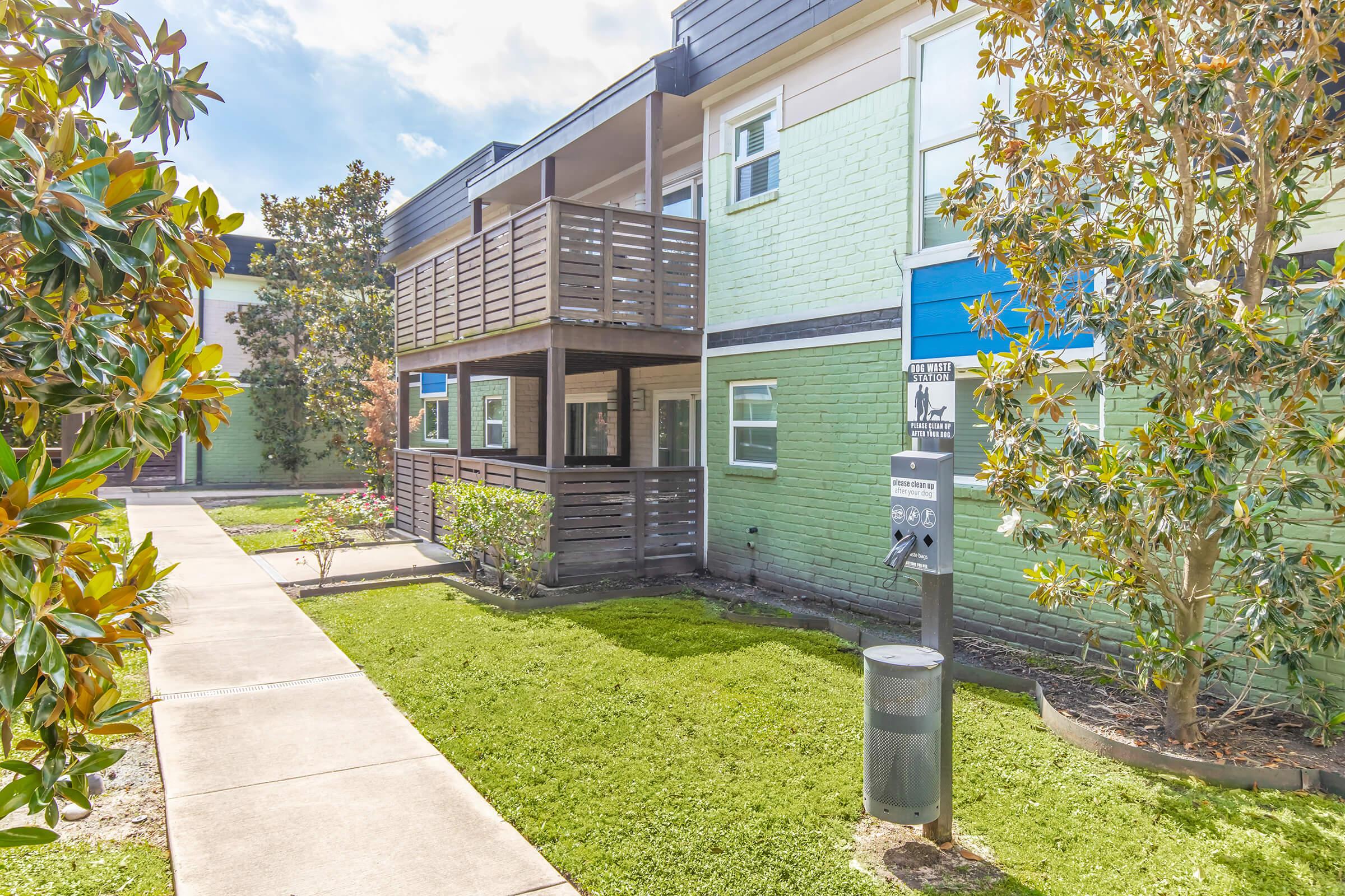 A well-maintained exterior of a two-story building with a green and white facade. Features include a balcony, landscaped garden with small bushes, and a paved walkway. There's also a parking meter visible in the foreground. The scene is bright and sunny, suggesting a pleasant environment.