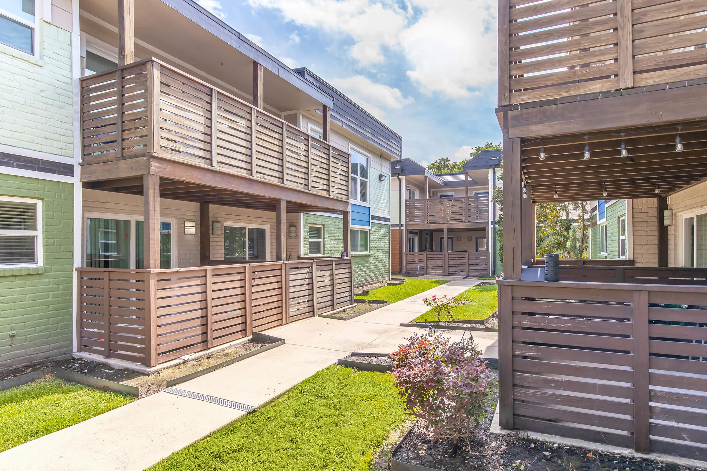 A view of a multi-unit residential building with multiple balconies, surrounded by well-maintained green lawns. Pathways connect the units, and the architecture features a mix of wooden and brick elements, reflecting a modern design. The sky is bright with fluffy clouds.