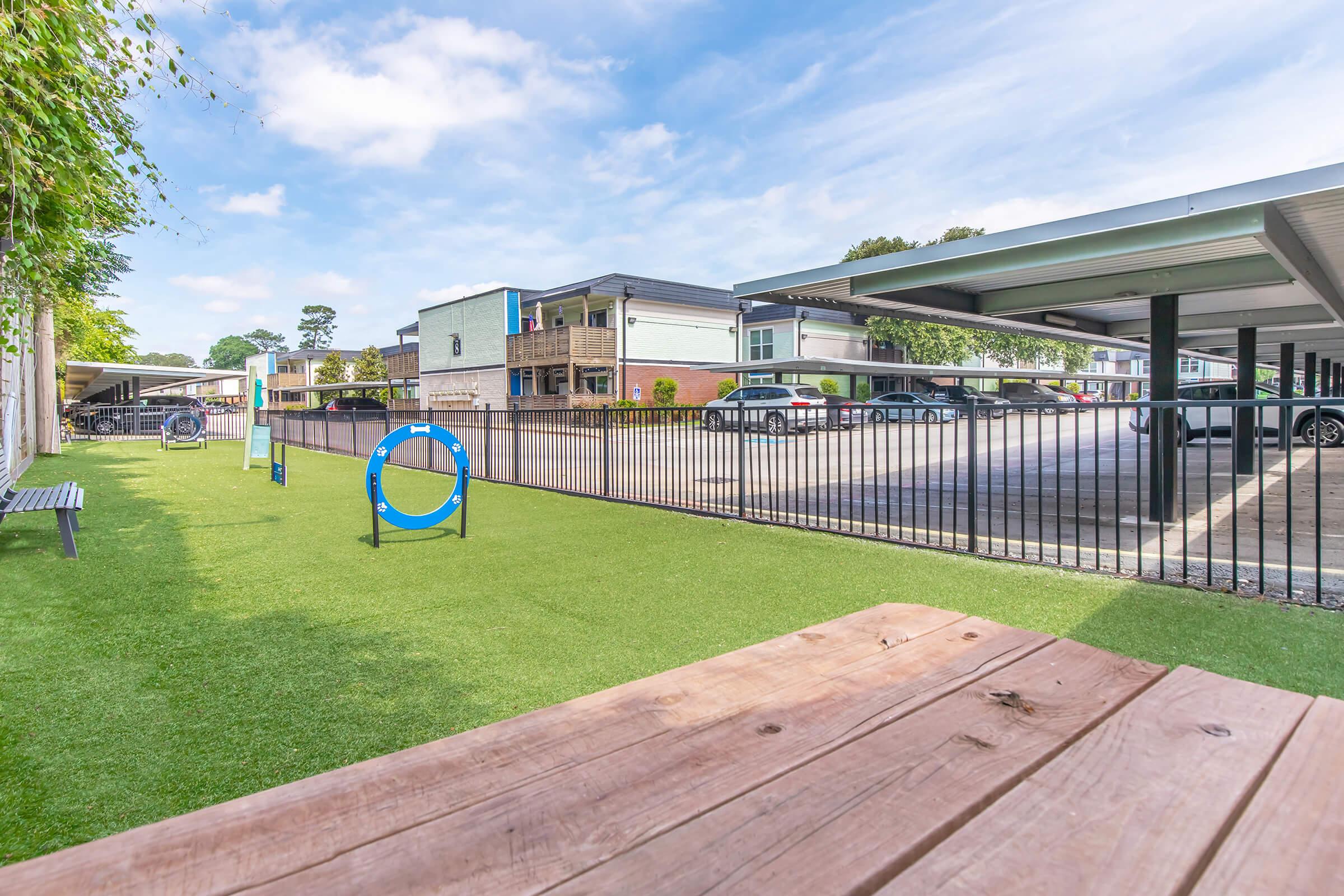 A view of a landscaped area featuring artificial grass, benches, and circular play equipment. In the background, there's a parking area with several cars and modern buildings. The sky is partly cloudy, creating a bright and inviting atmosphere.