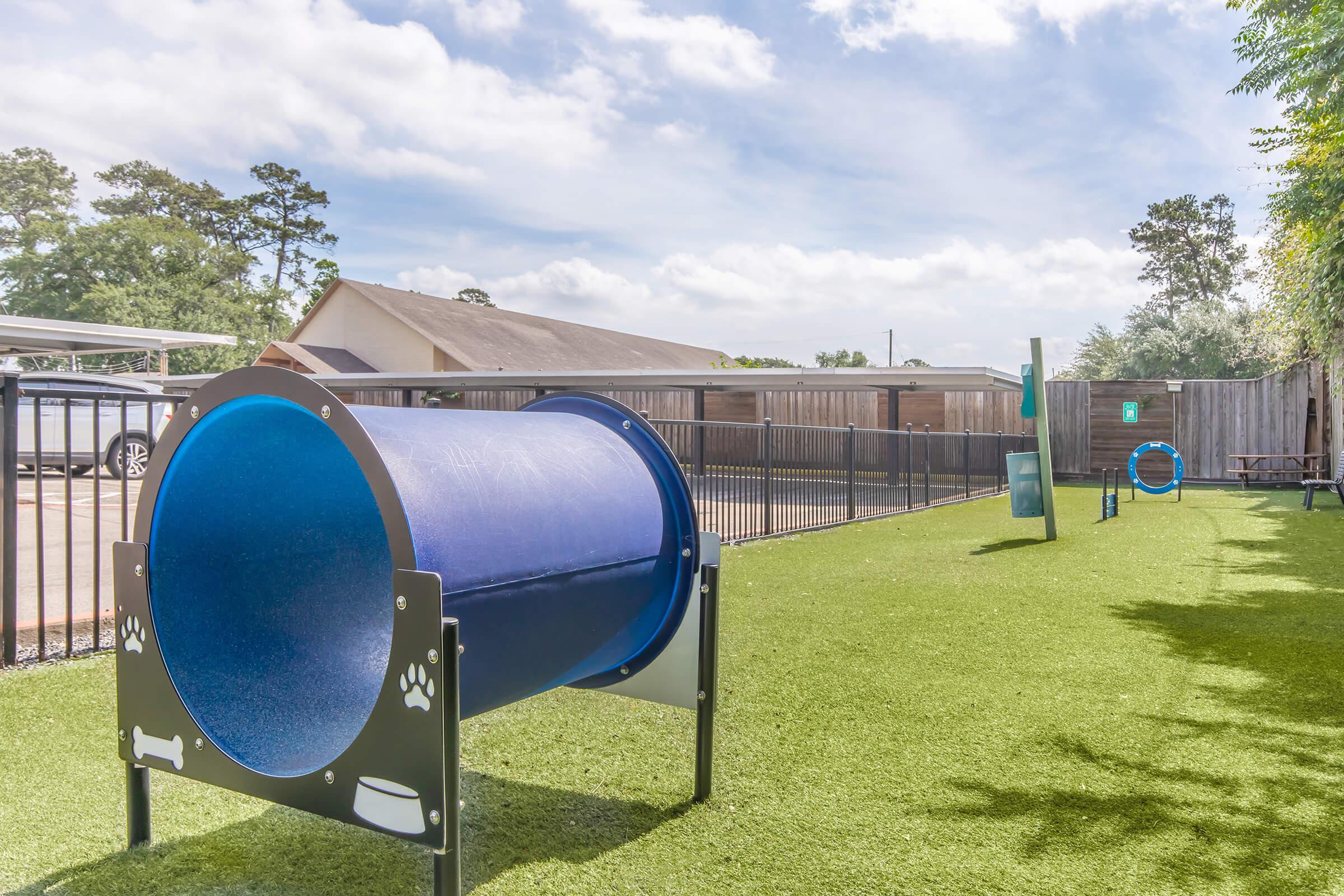 A dog agility course featuring a blue tunnel and a circular hoop on a grassy area. In the background, there are trees and a building. The scene is set under a partly cloudy sky, creating a cheerful outdoor environment for dogs to play and exercise.