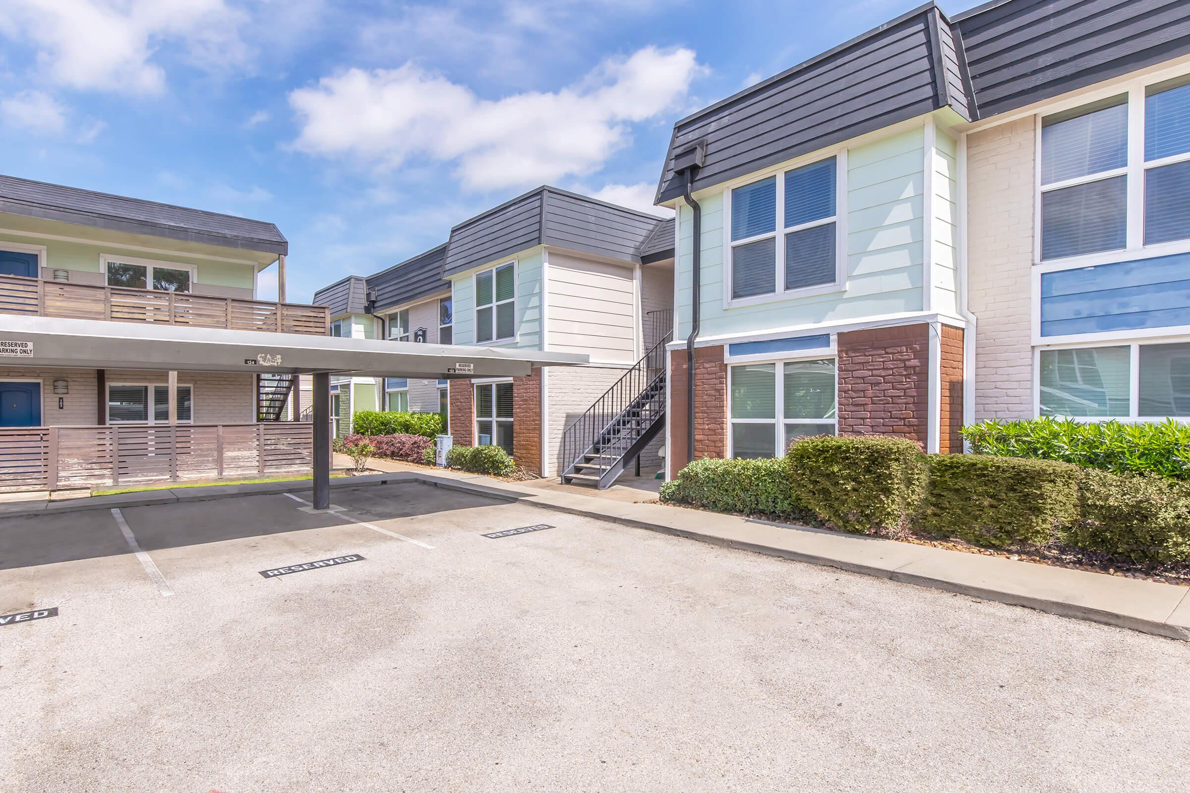Two-story townhouse-style apartments with a modern exterior. The building features a mix of light and dark siding, a covered parking area, and a well-maintained landscape. There are clear blue skies above, creating a bright and inviting atmosphere.