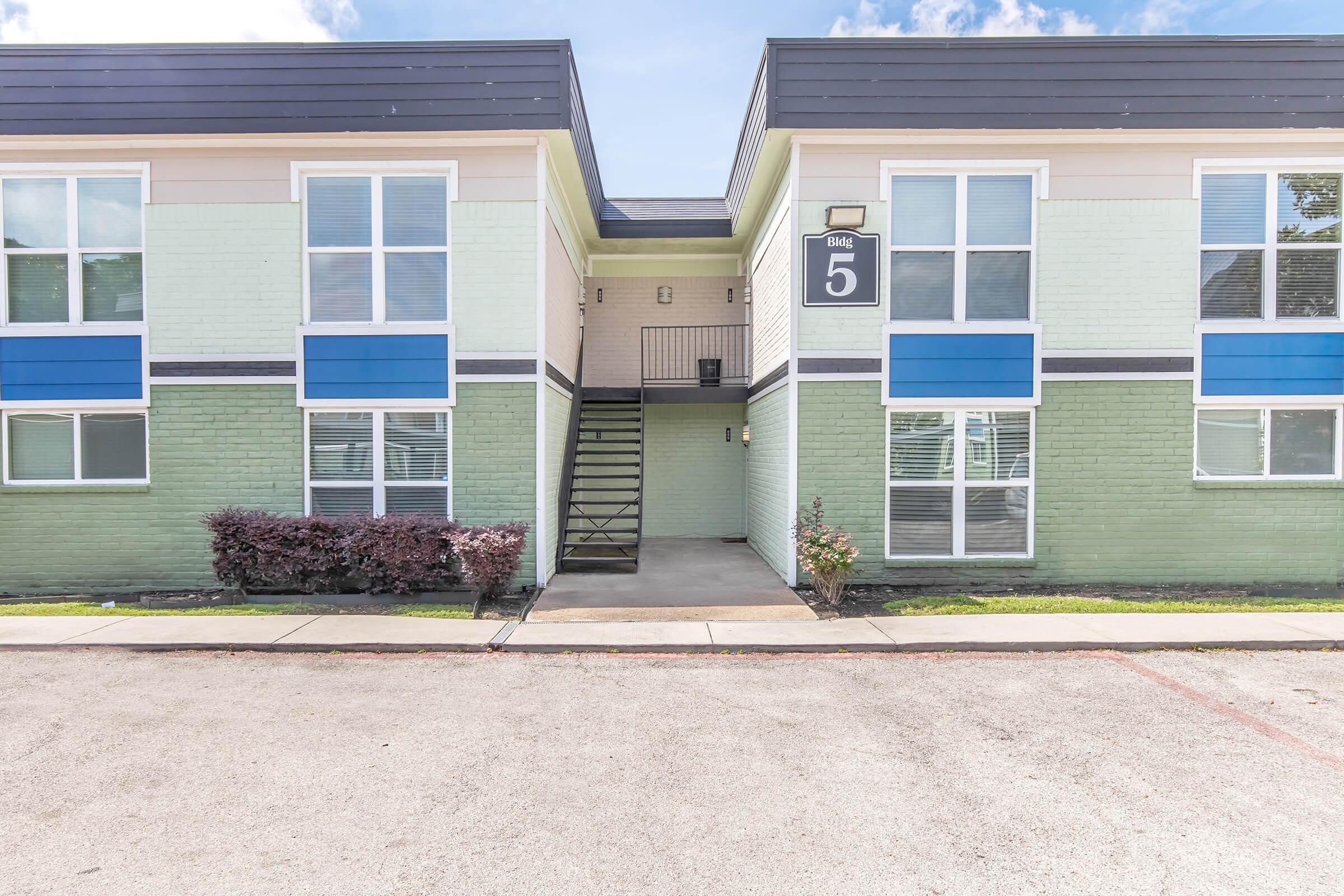 Exterior view of a two-story apartment building featuring green and white walls, large windows with blue accents, and a central staircase leading to the upper level. A small landscaped area with bushes is visible in front of the entrance. The building number is displayed as "5."