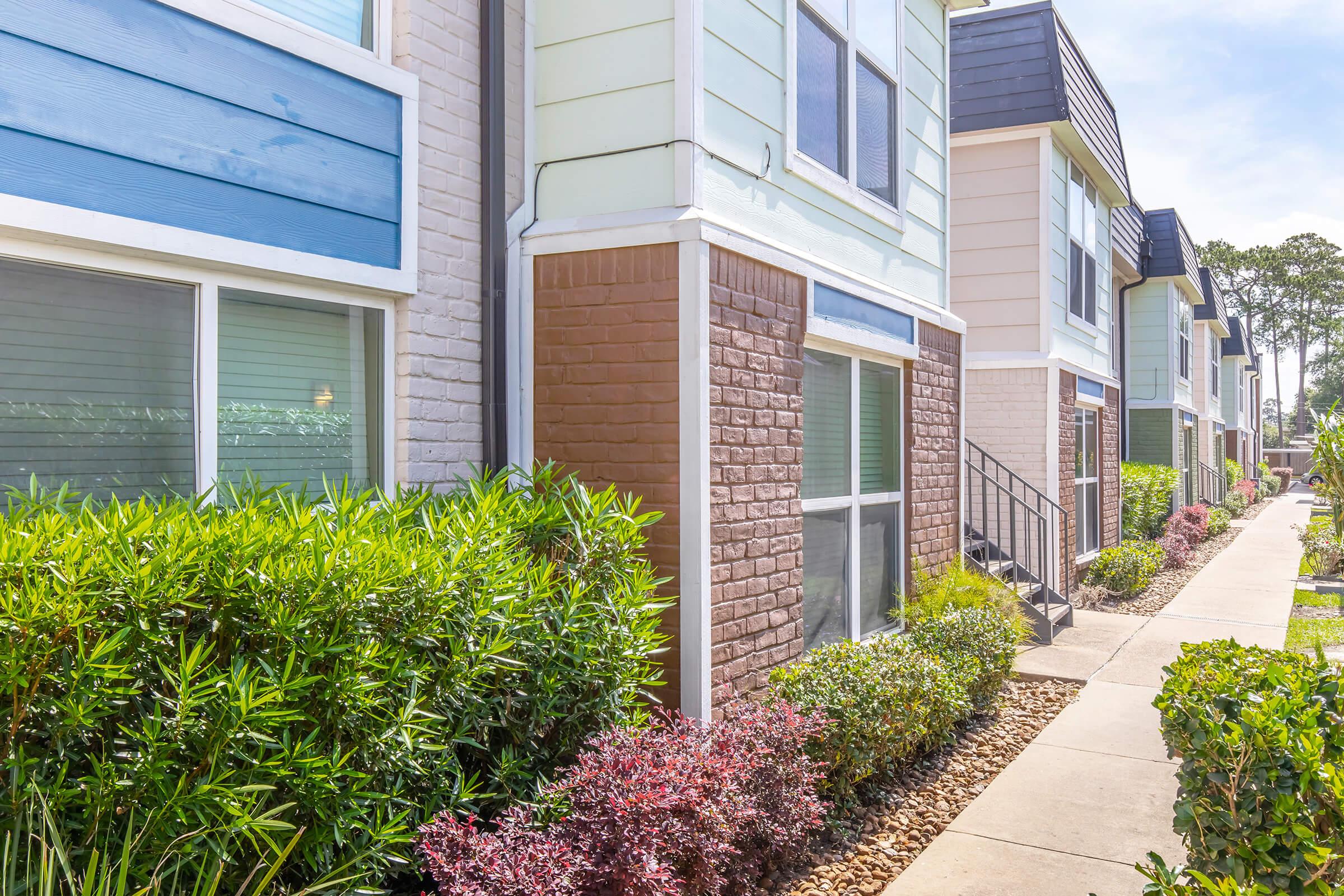 Row of townhouses with a mix of light blue, beige, and brown exteriors. The buildings are surrounded by well-kept landscaping featuring green shrubs and colorful flowers. A paved pathway runs alongside the homes, providing access to the entrances. Bright sky in the background suggests a sunny day.