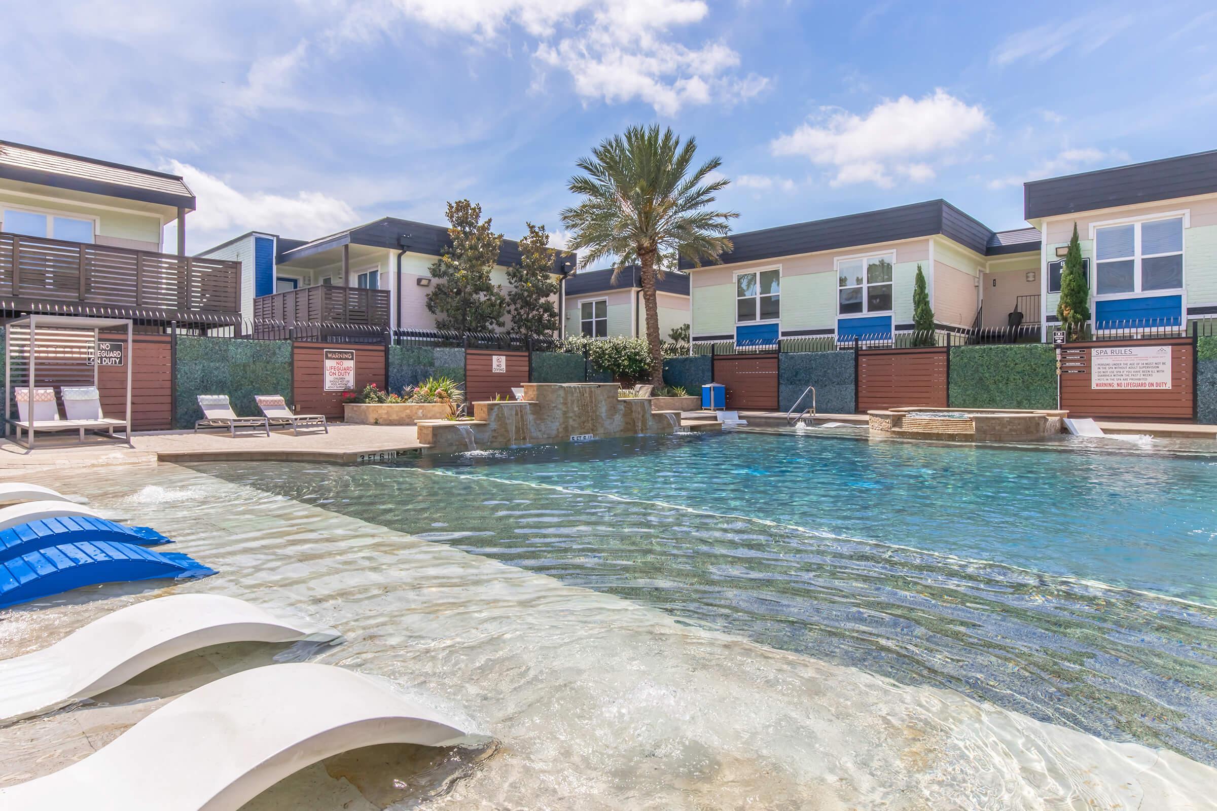 A tranquil swimming pool with a gentle waterfall feature, surrounded by lounge chairs and palm trees. Modern buildings with balconies are visible in the background under a blue sky with scattered clouds. The setting offers a relaxing atmosphere, perfect for recreation and leisure.