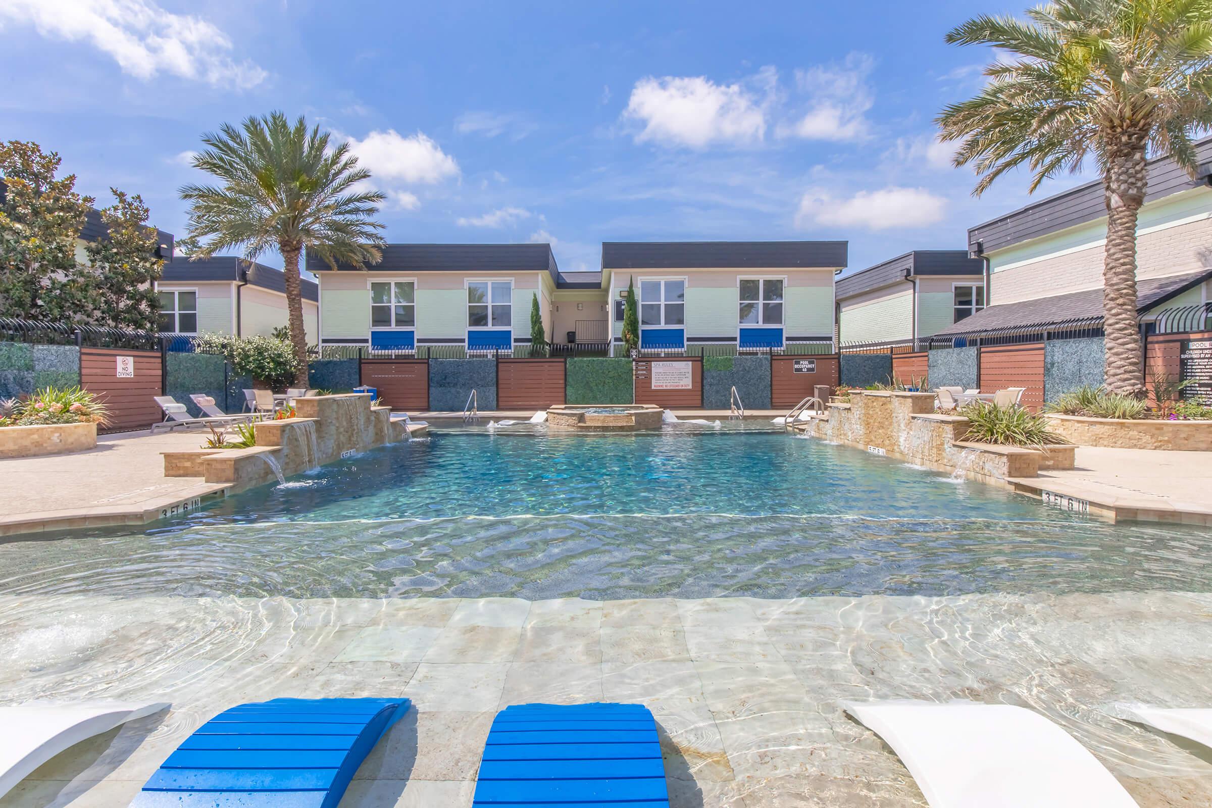 A serene swimming pool surrounded by tropical plants and palm trees, with lounge chairs on the poolside. In the background, modern buildings feature a contemporary design. The sky is clear with a few clouds, creating a bright and inviting atmosphere.