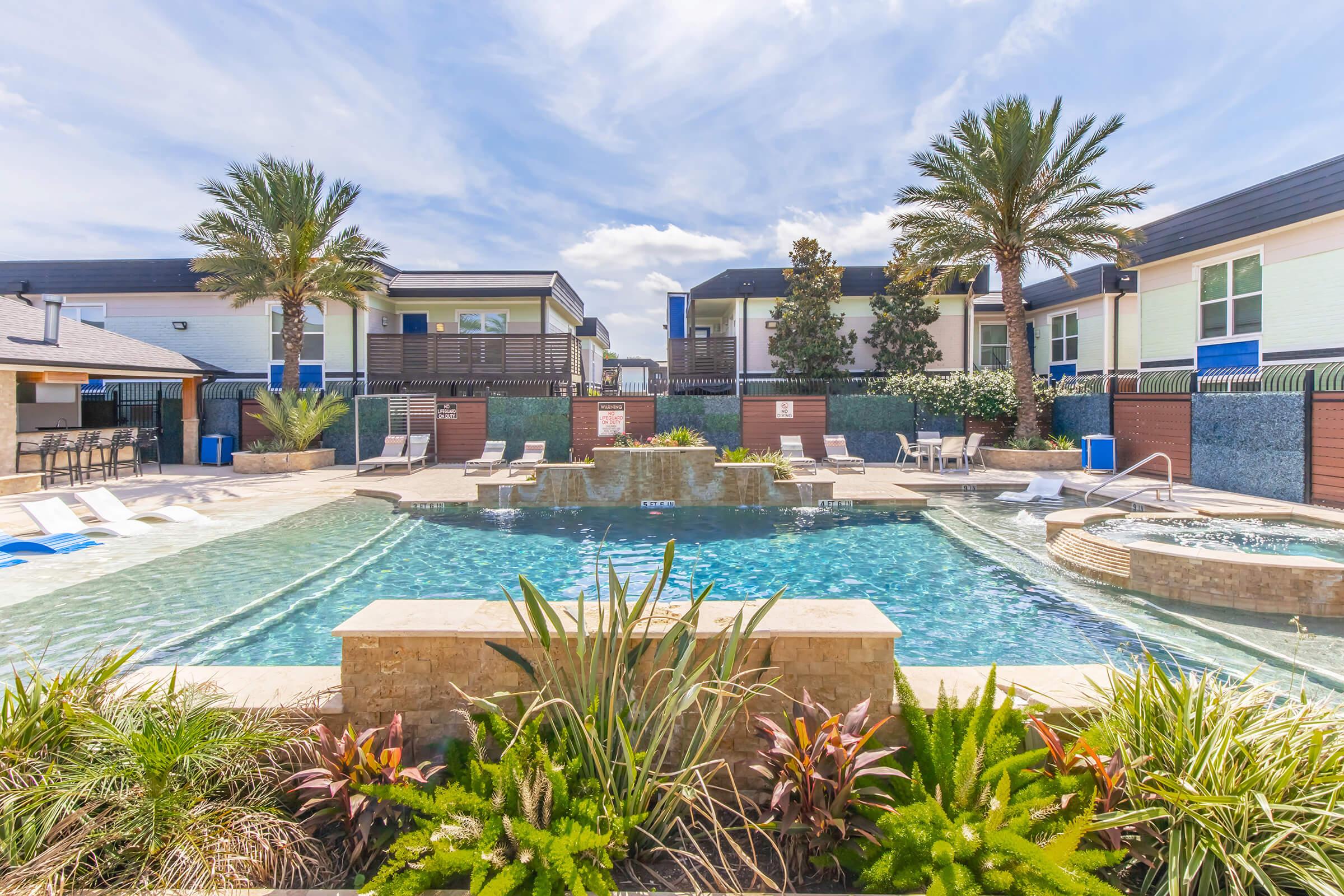 A vibrant outdoor pool area surrounded by landscaped greenery and palm trees. The pool features a central water fountain and is flanked by lounge chairs and a spa. Modern residential buildings are visible in the background under a partly cloudy sky.