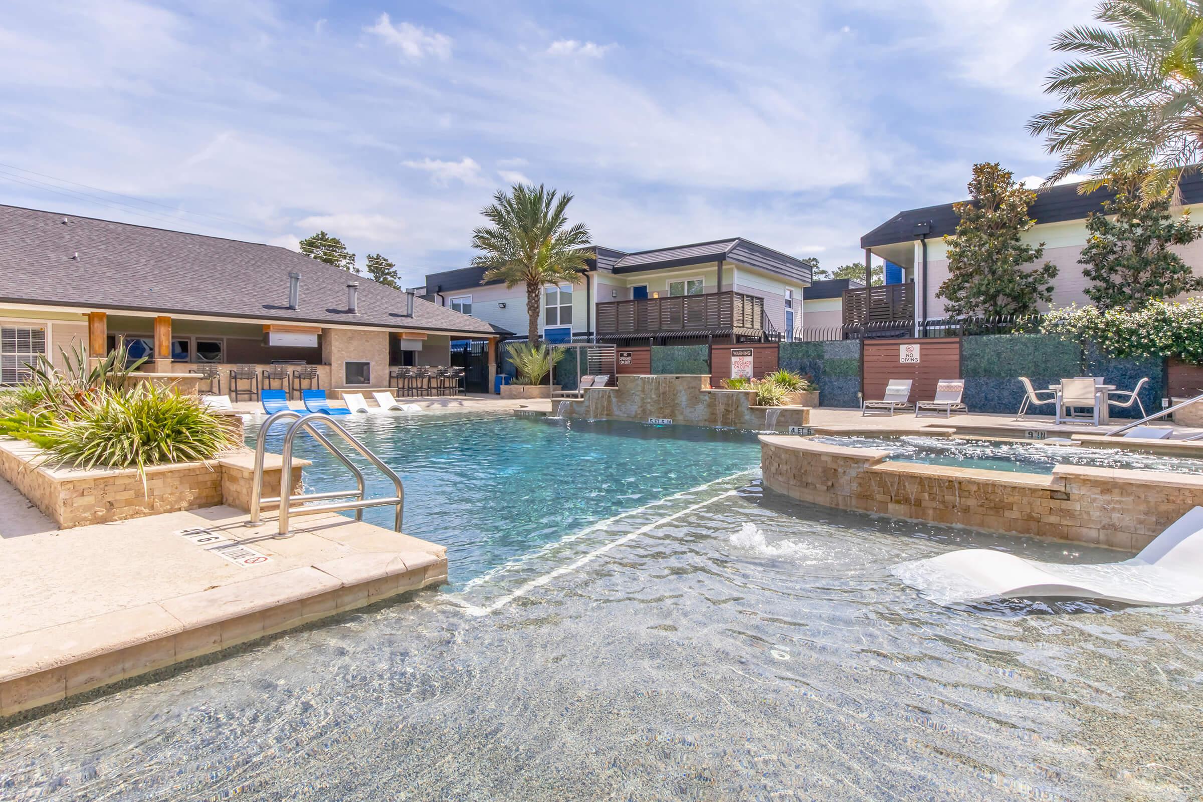 A sparkling swimming pool surrounded by palm trees and lounge chairs, with a shallow area featuring steps leading into the water. In the background, there are modern buildings and a sunny sky, creating a relaxing atmosphere. The pool area is well-maintained and inviting.