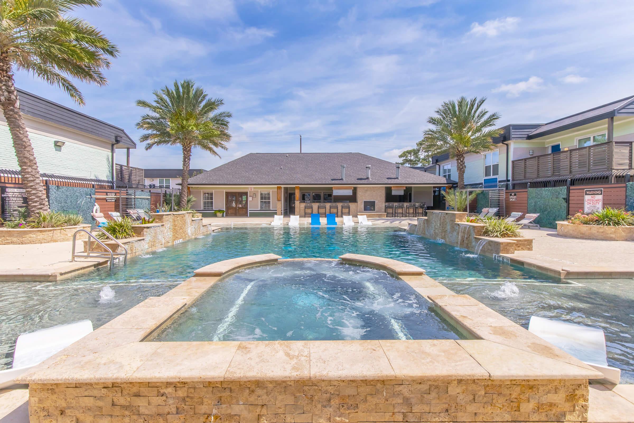 A sparkling swimming pool surrounded by palm trees, lounge chairs, and landscaped greenery. In the background, a building with a dark roof and outdoor seating area can be seen. The sky is bright and clear, adding to the inviting atmosphere of the outdoor space.