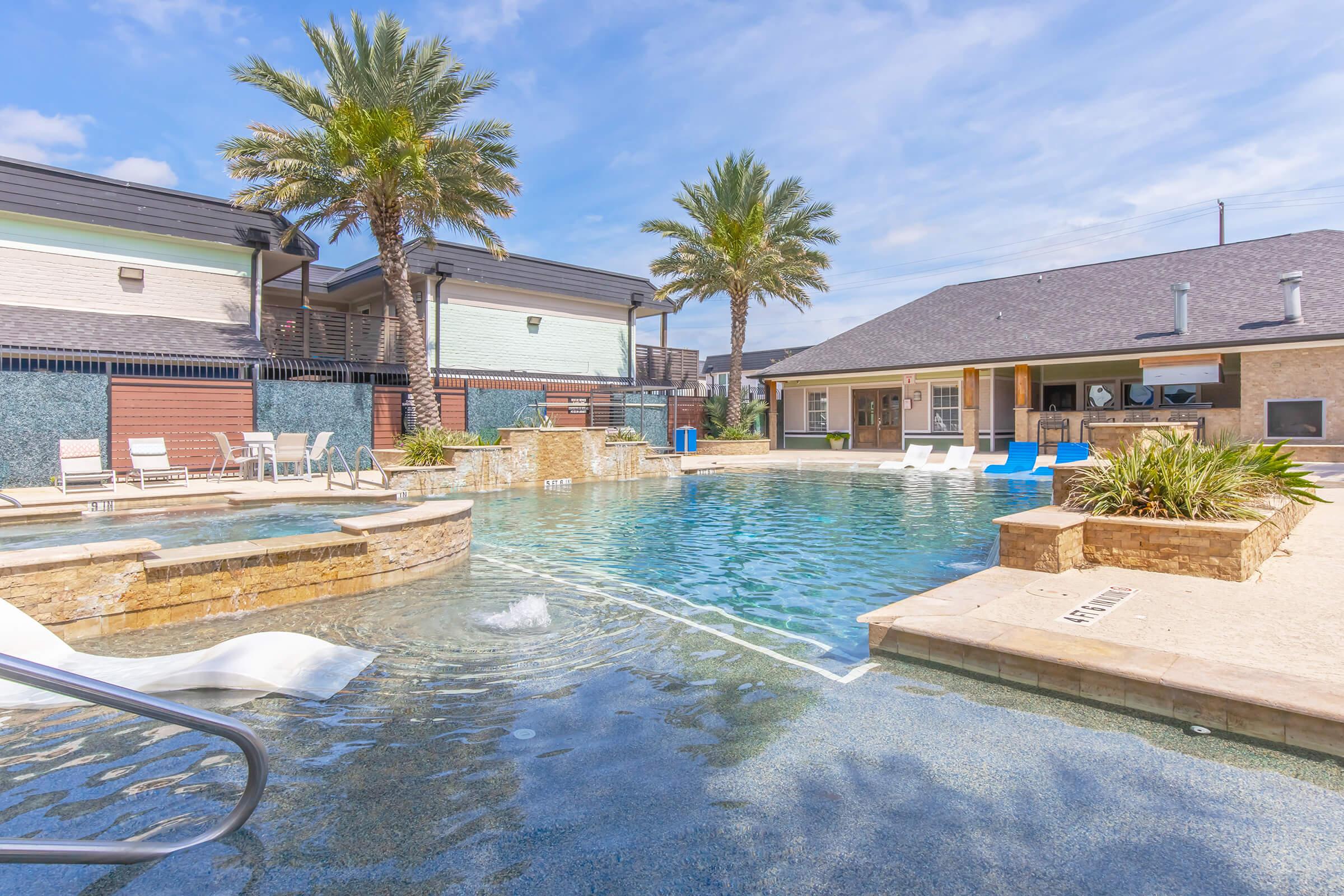 A sunny outdoor pool area surrounded by palm trees, featuring a clear blue pool with a stone deck, lounge chairs, and a nearby clubhouse. The sky is bright with a few clouds, creating a relaxing atmosphere perfect for swimming and lounging.