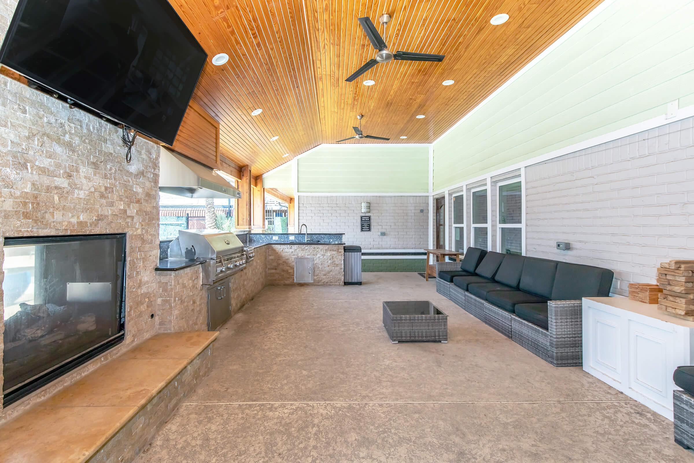Spacious outdoor lounge area with a ceiling fan and wooden accents, featuring a built-in grill, television, and a stone fireplace. There are modern furniture pieces, including a dark wicker sofa and a white cabinet, all under a covered patio with ample natural light. Perfect for entertaining or relaxing.
