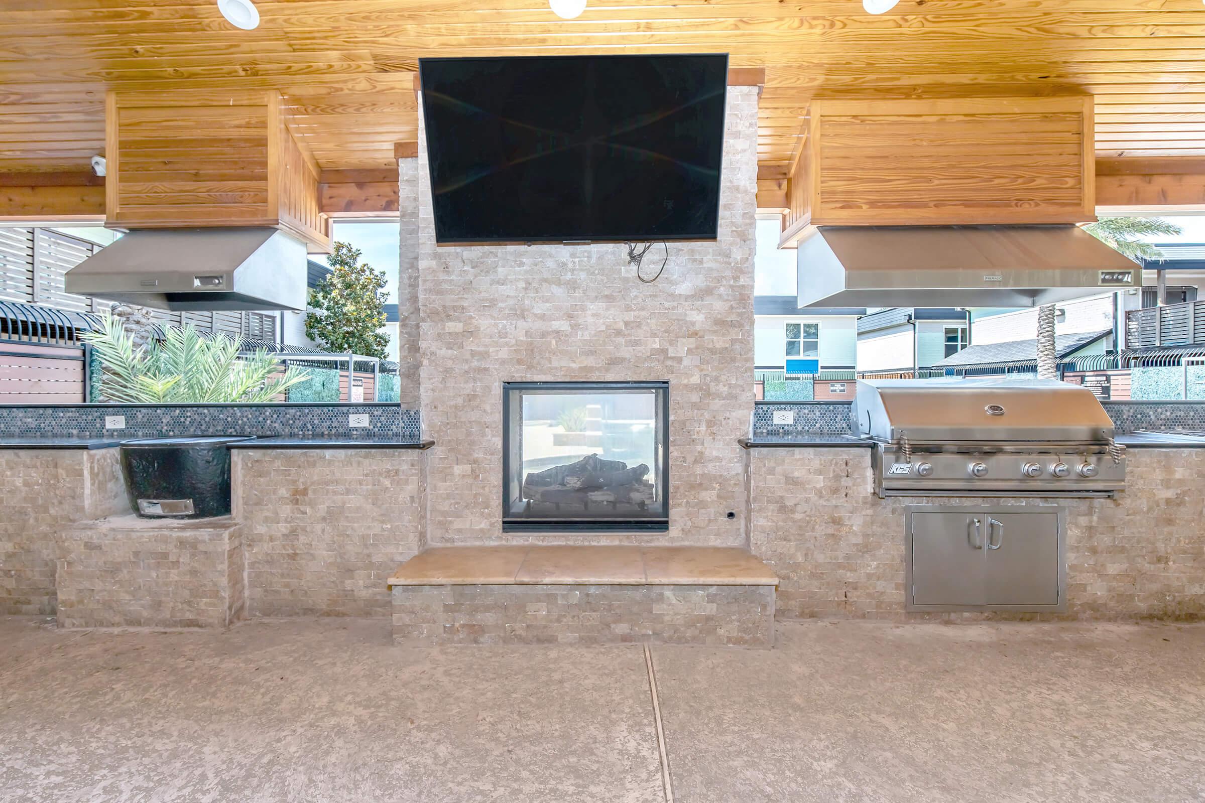 Outdoor kitchen area featuring a stone fireplace with a modern TV above, a built-in grill with stainless steel cabinets, and a bar-style countertop. The space is covered by a wooden ceiling and surrounded by greenery, creating a stylish and inviting atmosphere for outdoor cooking and entertaining.